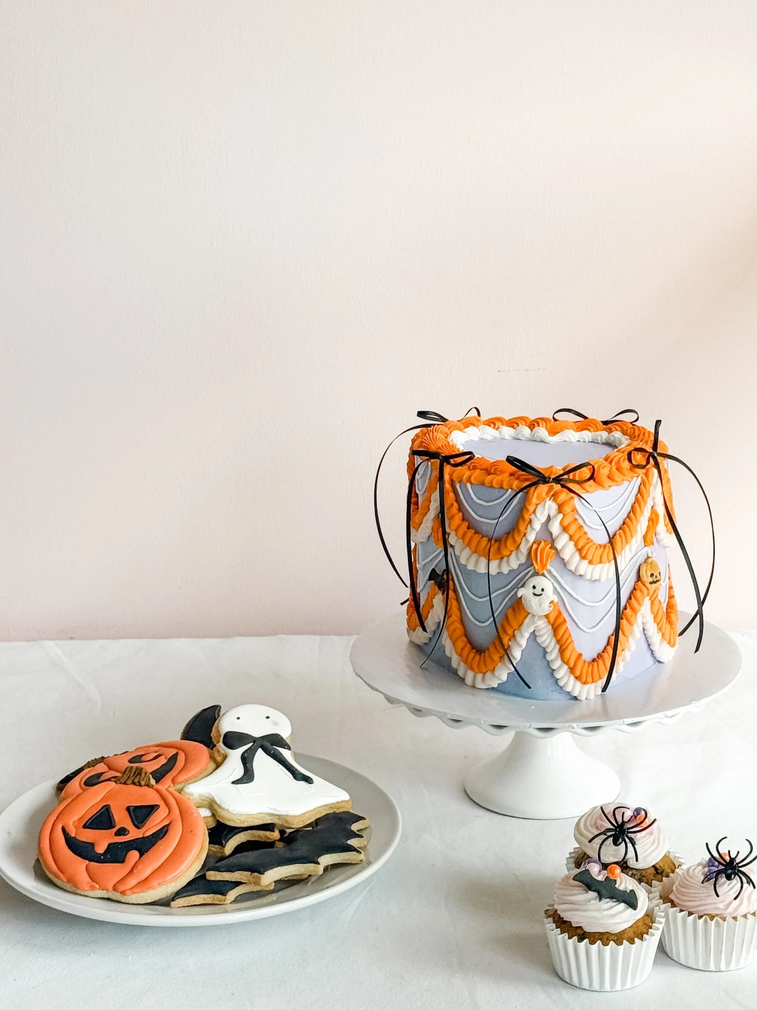 Lavender Halloween cake with orange and white vintage buttercream piping, black bows, ghosts and pumpkins. Along with three festive halloween cupcakes and decorated halloween sugar cookies.