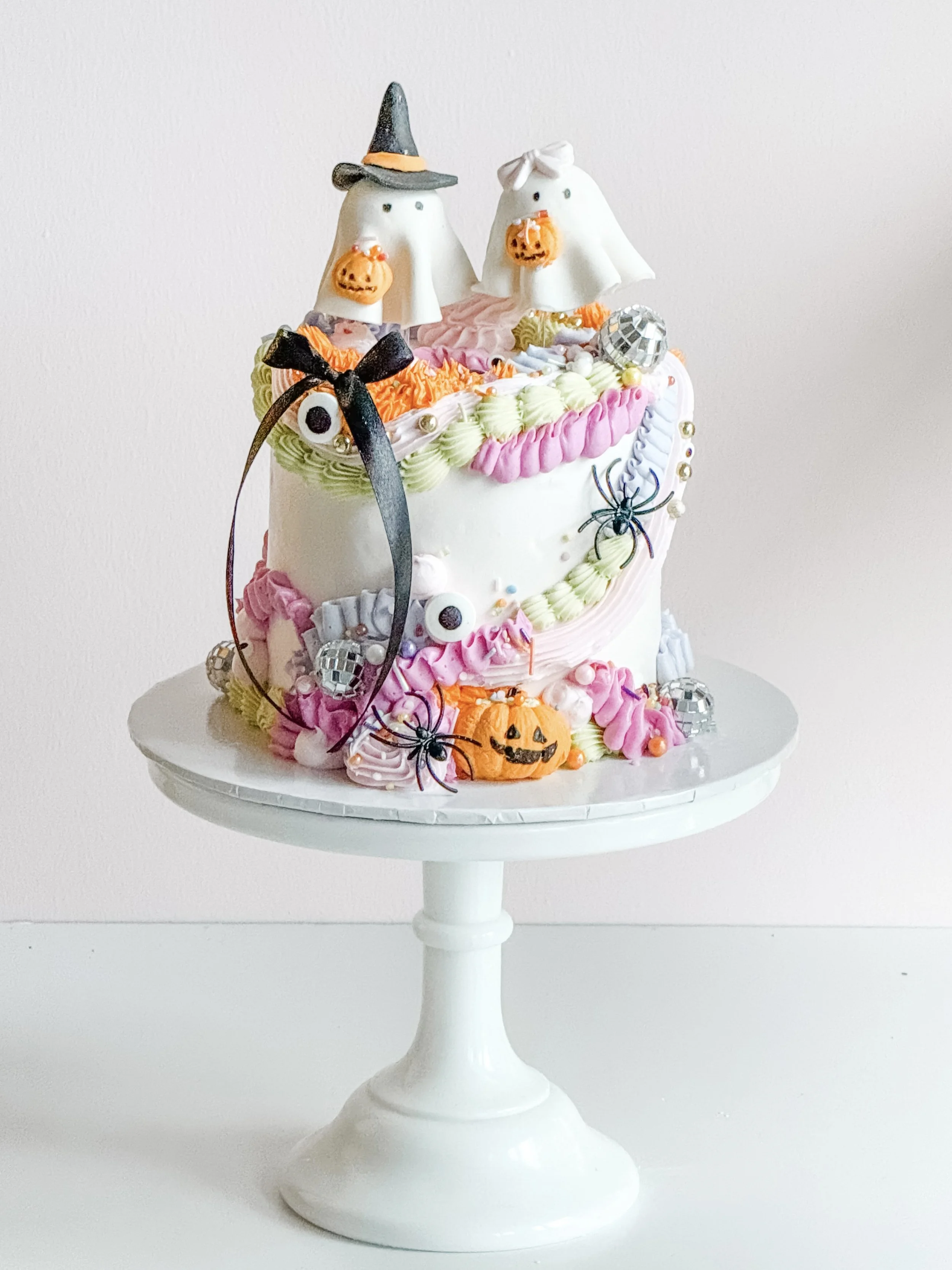 Boo's & Bows Halloween cake by Sweet Stacy's Palm Beach with two white ghost toppers, pastel buttercream piping, black bows, mini disco balls and pumpkins