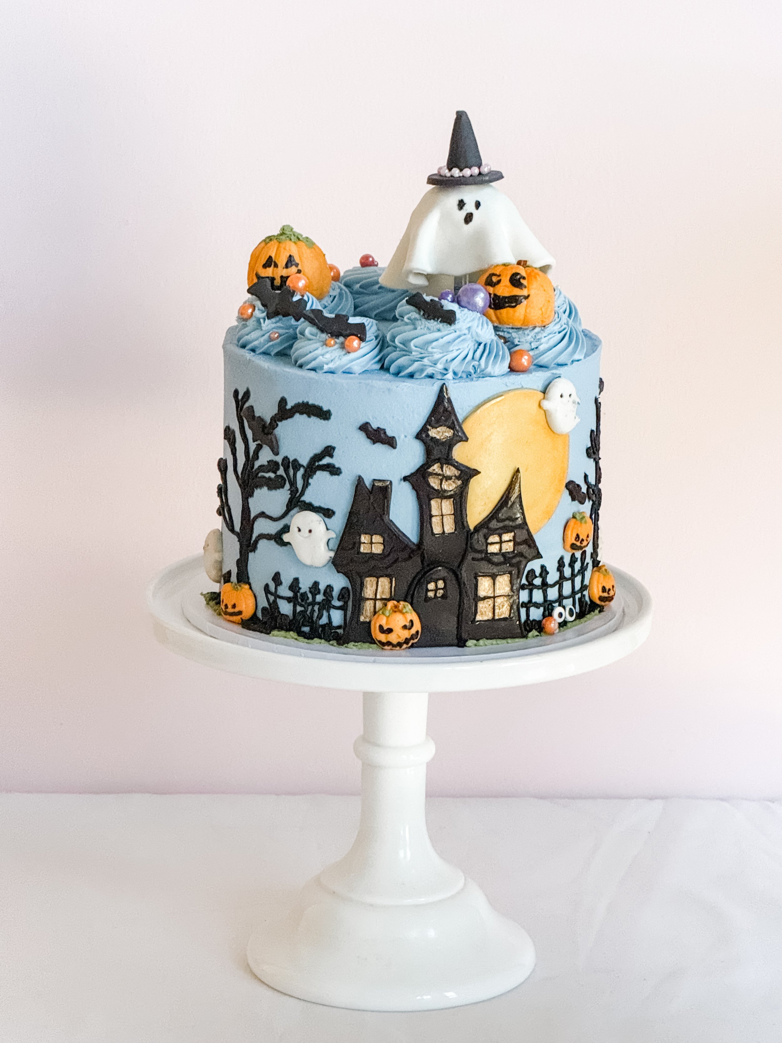 Blue Haunted Soirée Halloween cake by Sweet Stacy's Palm Beach featuring a black haunted mansion, golden moon, pumpkins, bats, and a ghost wearing a witch hat.