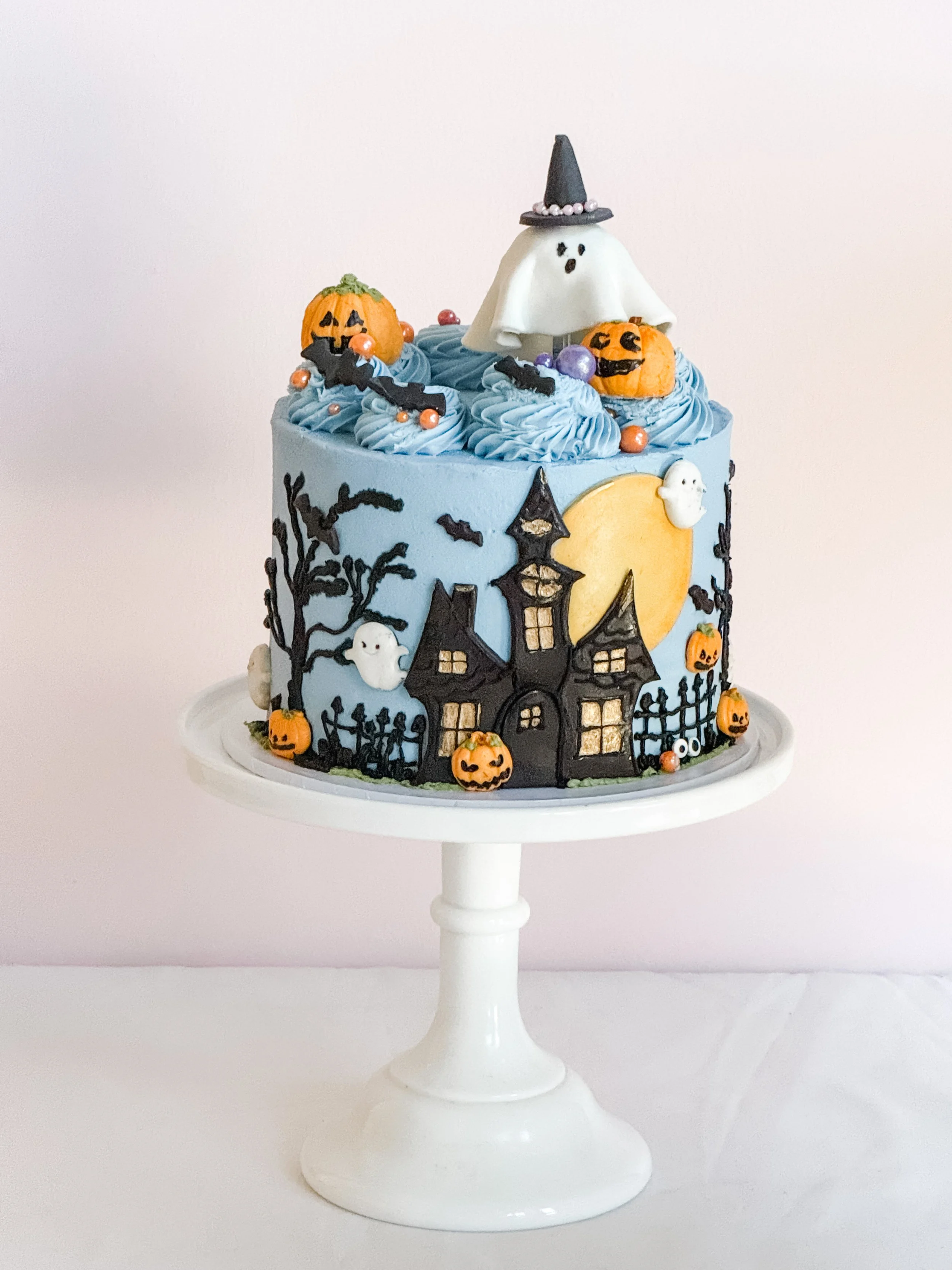 Blue Haunted Soirée Halloween cake by Sweet Stacy's Palm Beach featuring a spooky black haunted house, glowing golden moon, pumpkins, flying bats, and a ghost topper.