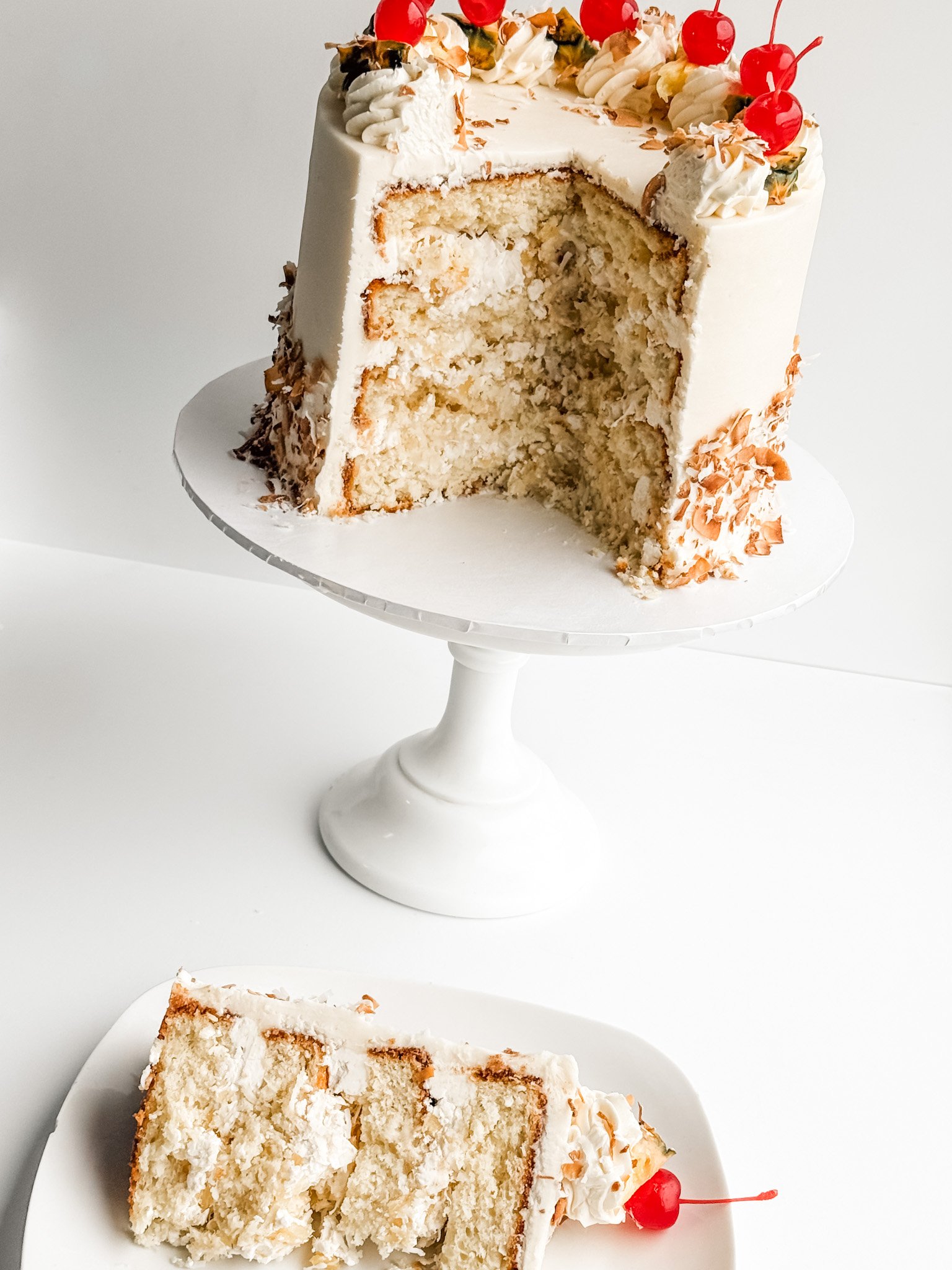 pina colada cake showing the layers and decorated with toasted coconut, pineapple, and cherries and a slice of the cake on a plate