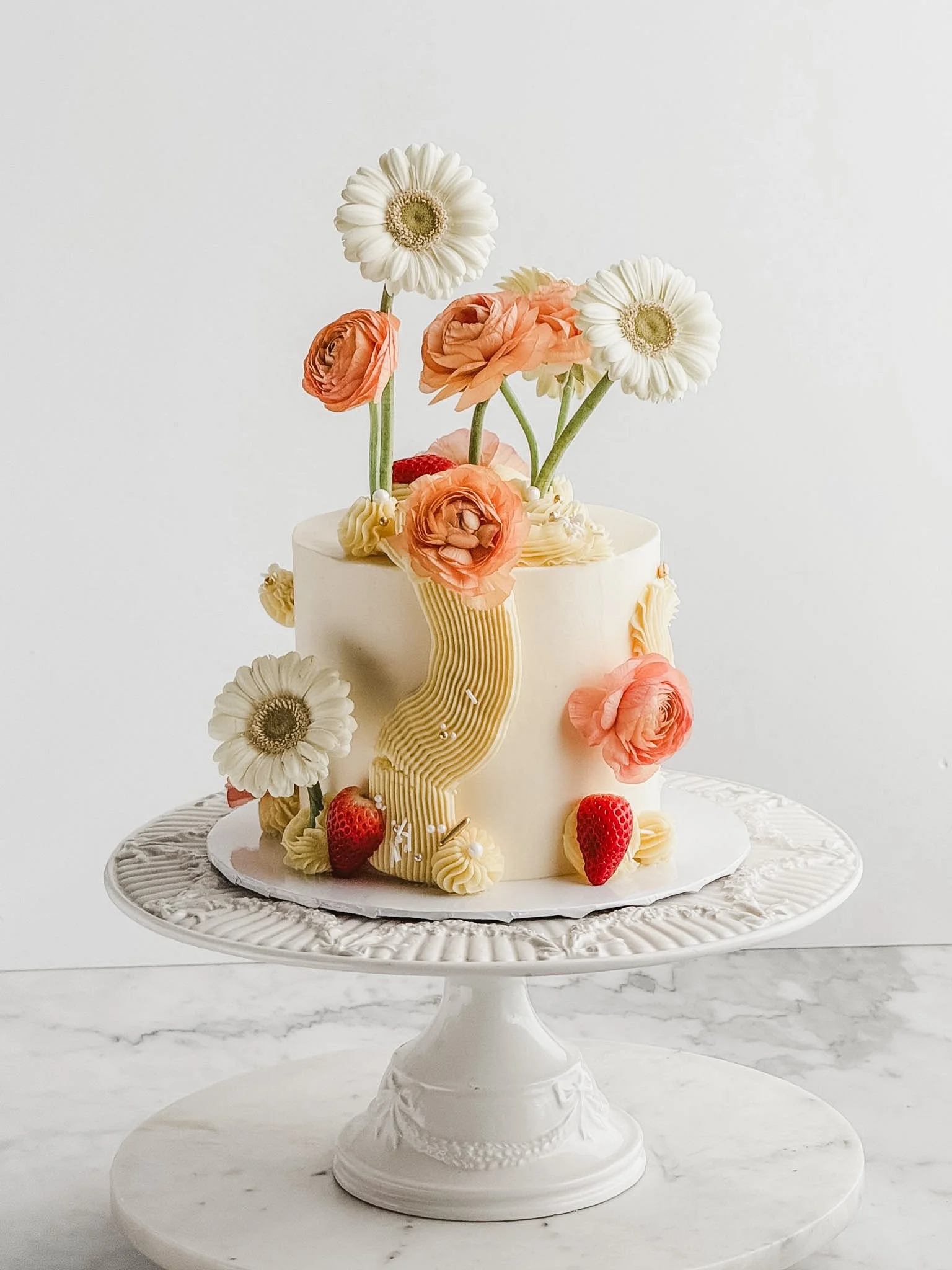 beautifully decorated spring cake with a yellow geometric pattern design and an array of florals in white and peach sitting on top of a cake stand