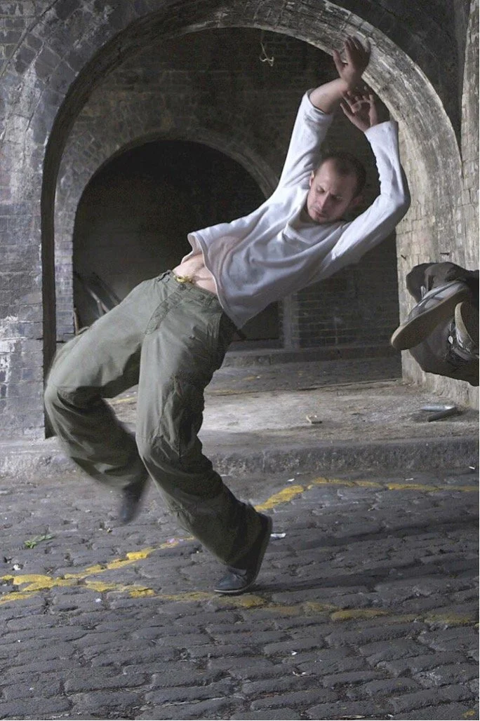 A person, Leon Baugh, in a white long-sleeve shirt and baggy green pants performs an urban dance move in a gritty, archway tunnel setting