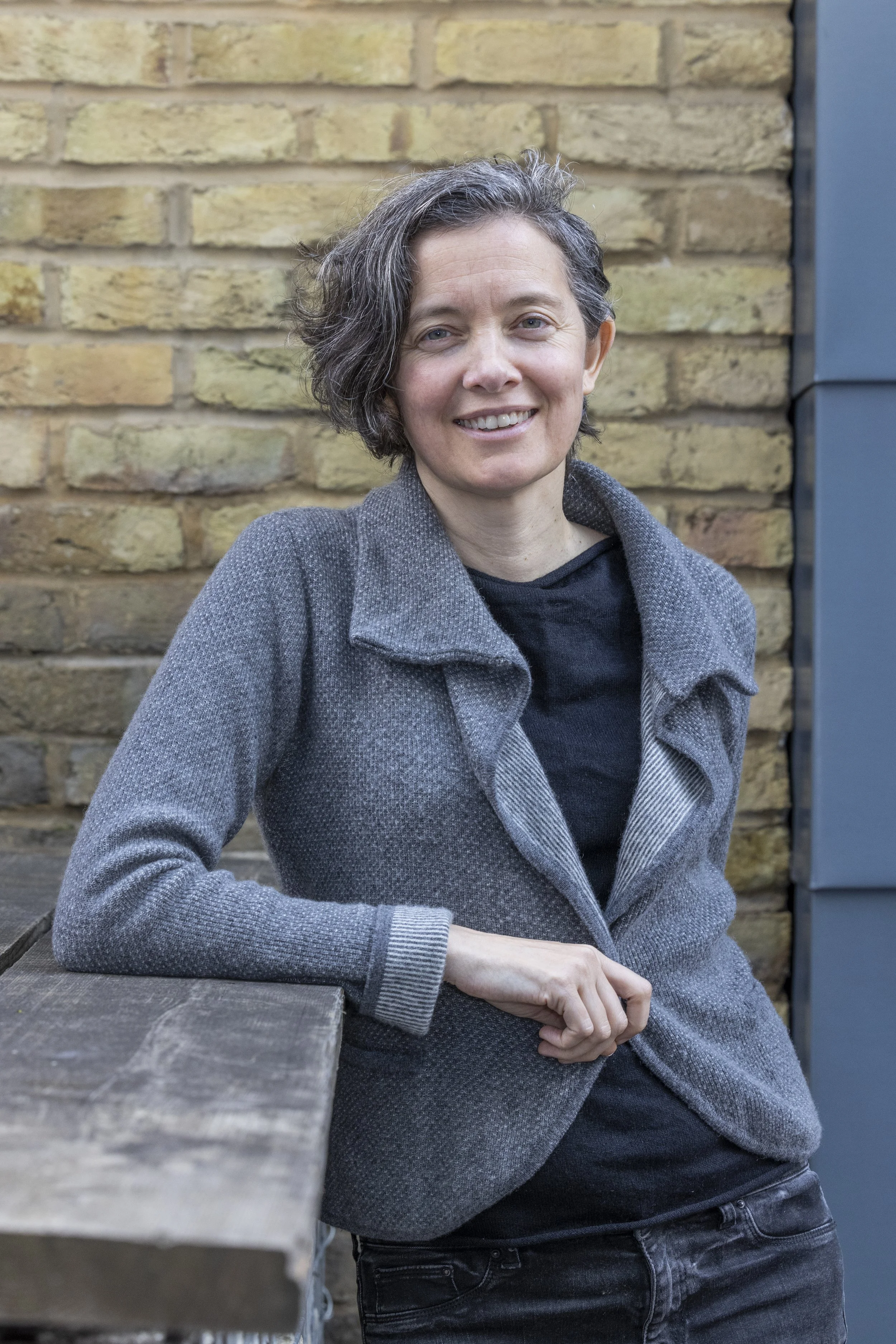 A person, Sacha Wares , in a cozy gray cardigan leans casually against a wooden railing, set against a backdrop of textured brick walls.