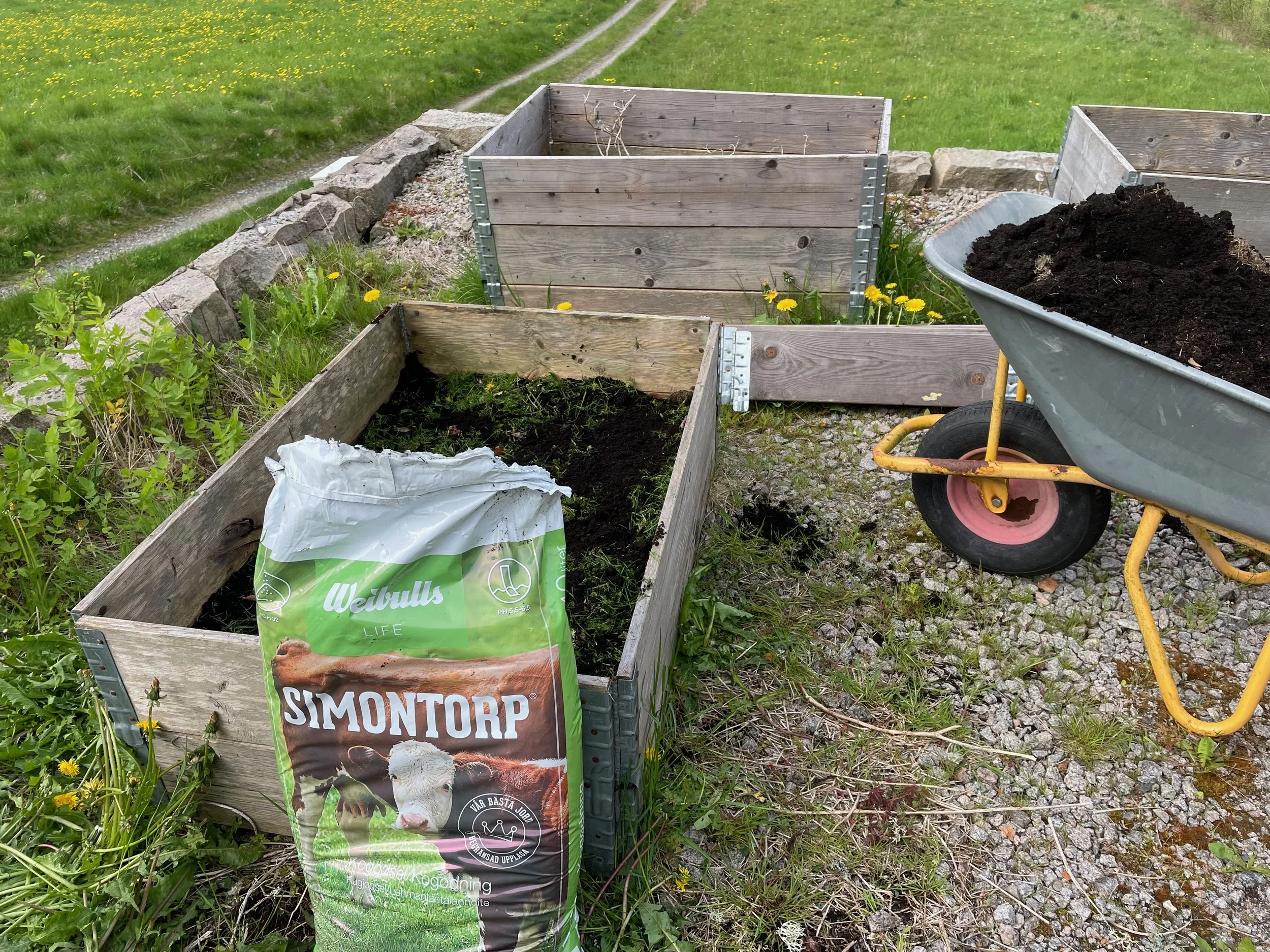 Empty wooden garden beds, a bag of soil labeled 'SimTorp', a wheelbarrow filled with dark soil, and some green plants and yellow flowers surrounding the beds outdoors.