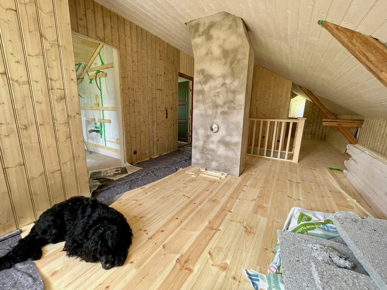 A black dog lying on a wood floor in an unfinished room with wooden panel walls and sloped ceiling, construction materials, and a partially built staircase.