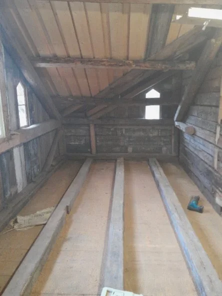 Inside of an unfinished attic or loft space with exposed wooden beams and walls, with three parallel floor joists laid out on the floor, and a cordless drill on the right side.