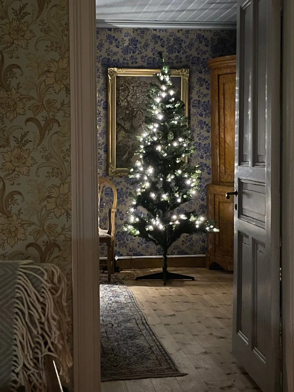 A decorated Christmas tree with white lights in a room with floral-patterned wallpaper and wooden furniture.