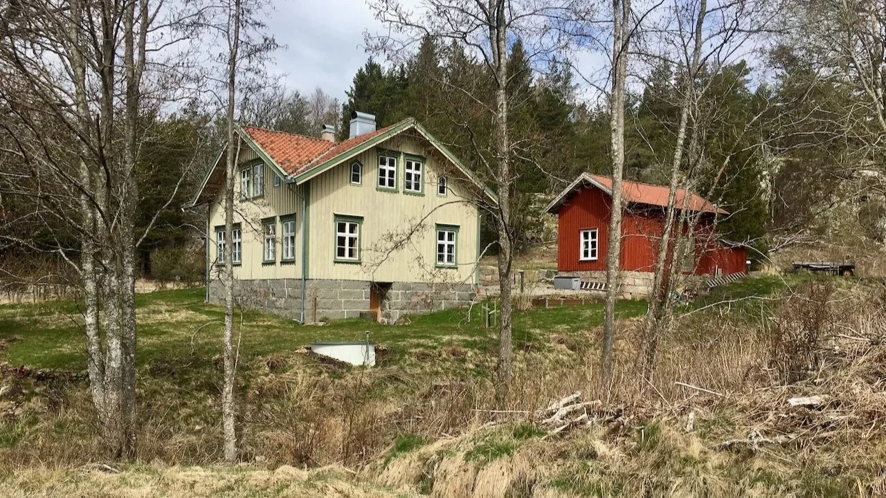 Two houses in a wooded area, one painted light yellow with a red tile roof, the other painted red with a metal roof, surrounded by bare trees and grass.