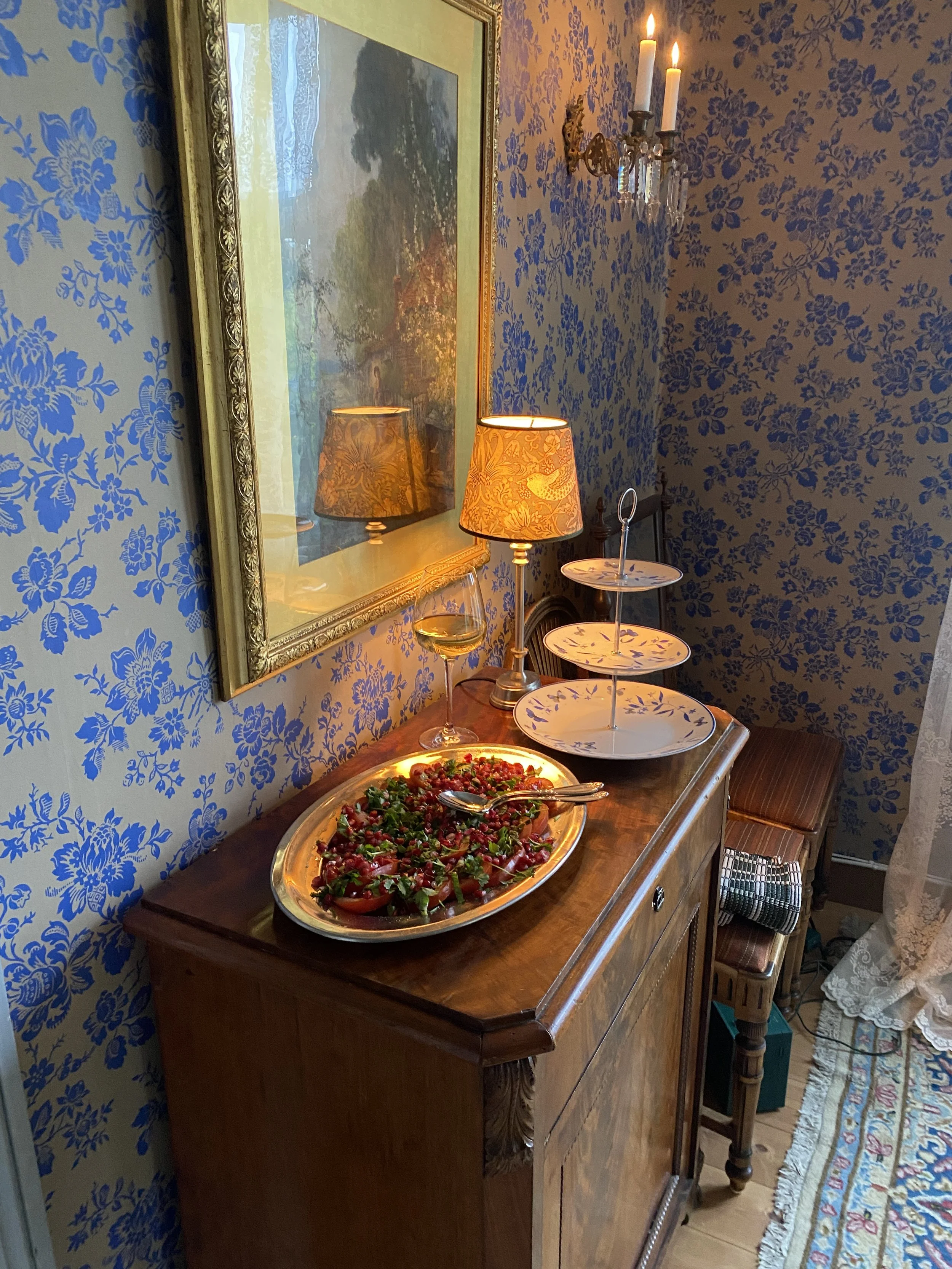 A wooden sideboard with a large oval platter of salad, a glass of white wine, two lamps with decorated shades, and a three-tiered serving stand. There is framed artwork on a blue floral wallpapered wall, a small bench with a checkered cloth, and a ca