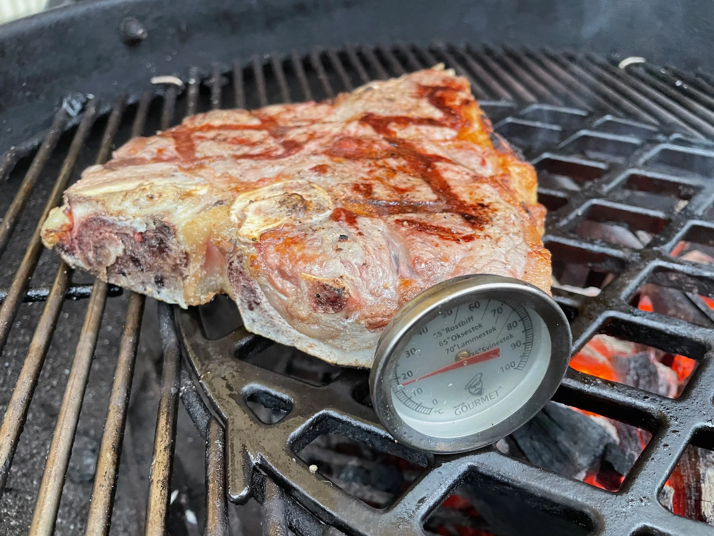 A thick T-bone steak cooking on a grill with a meat thermometer inserted to check its temperature.