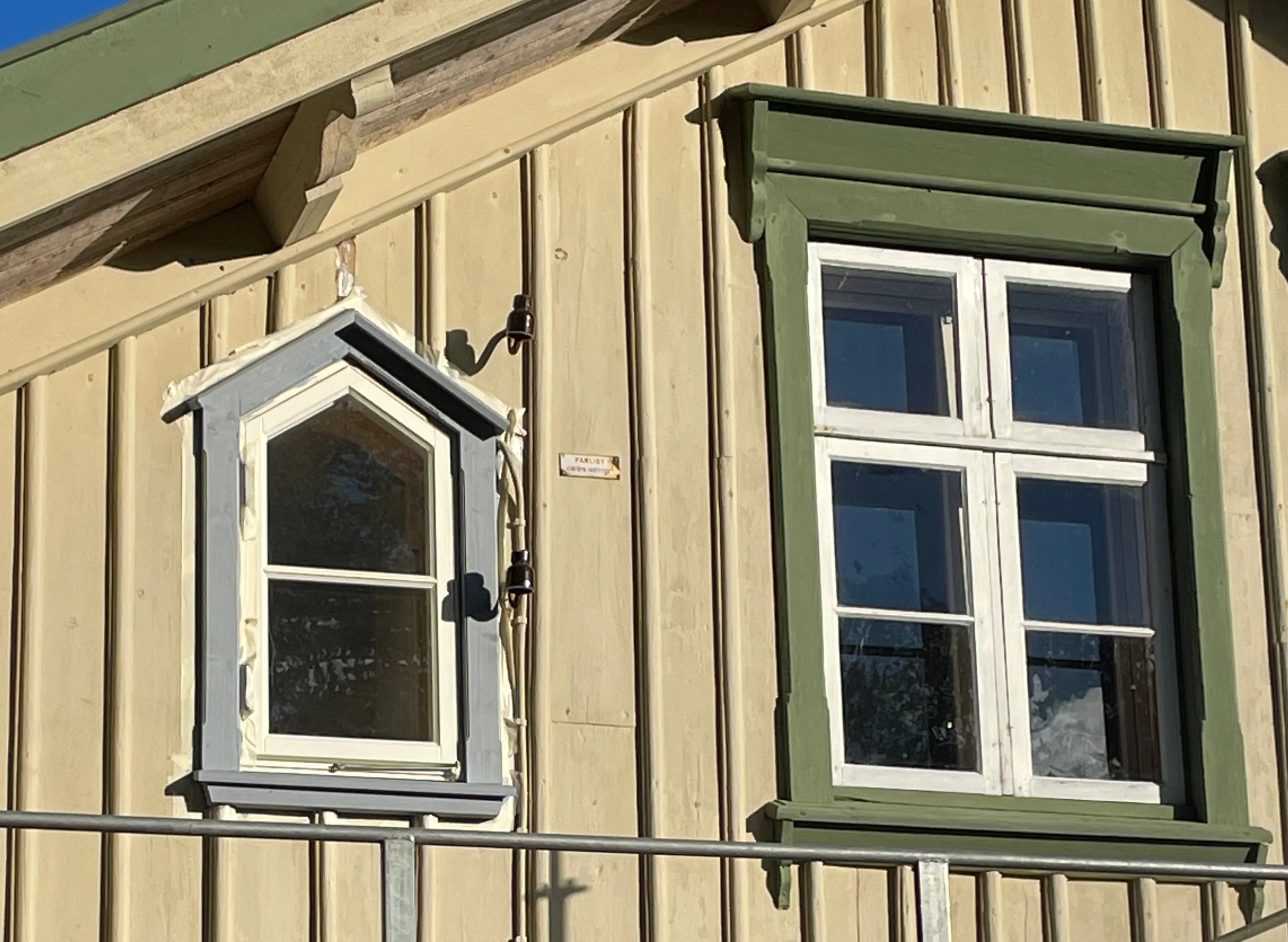 Close-up of two window sections on a wooden house wall, one with white trim and a pointed top, the other with green trim and four panes, with a metal railing in front.