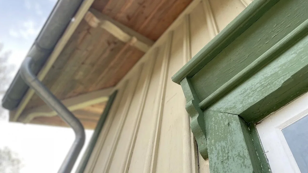 Close-up of a green wooden window frame with detailed molding, part of a beige siding wall under a wooden roof with metal gutter.
