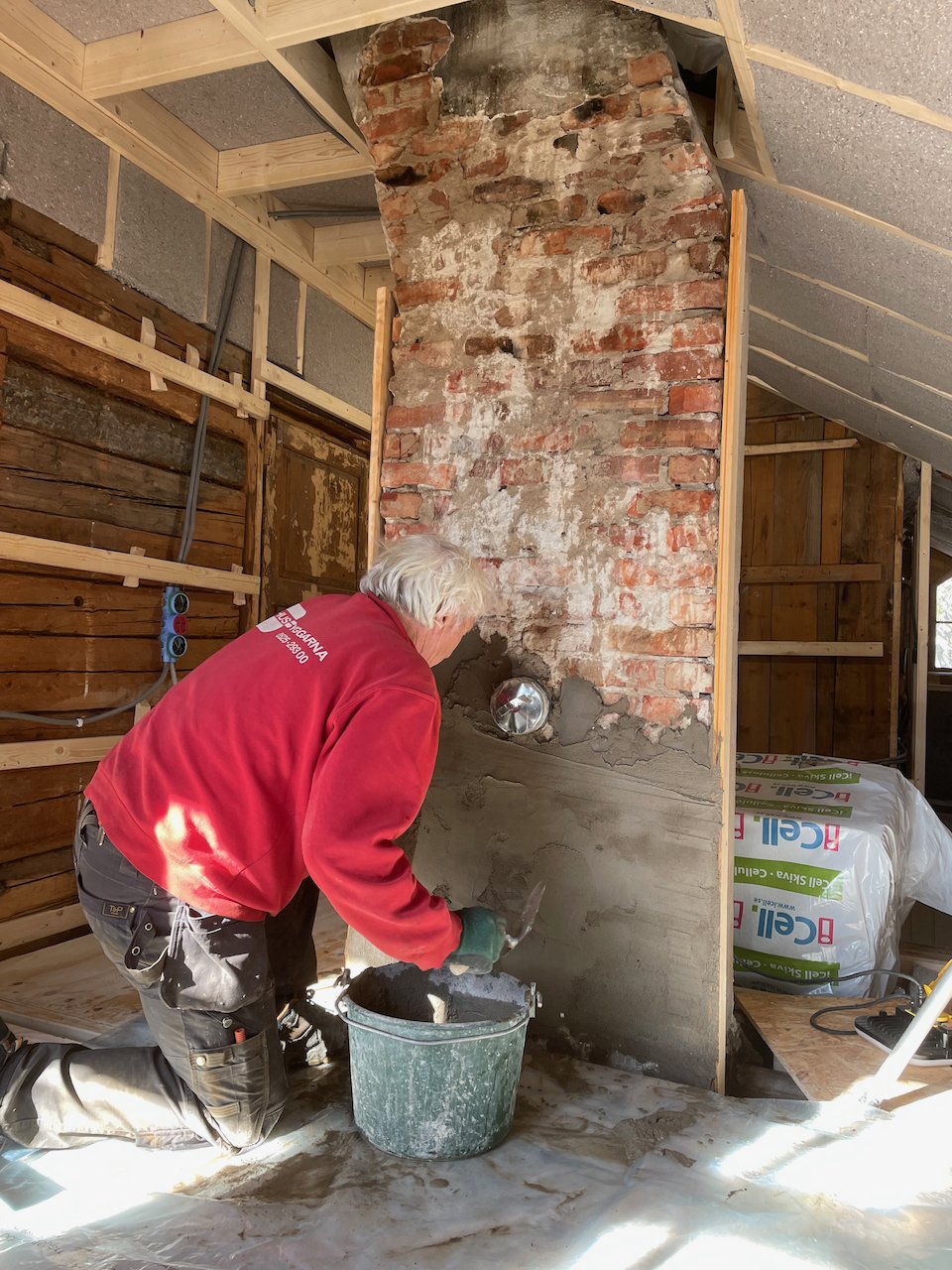 A person with gray hair working on a brick chimney in an unfinished room, mixing mortar in a bucket with a trowel.