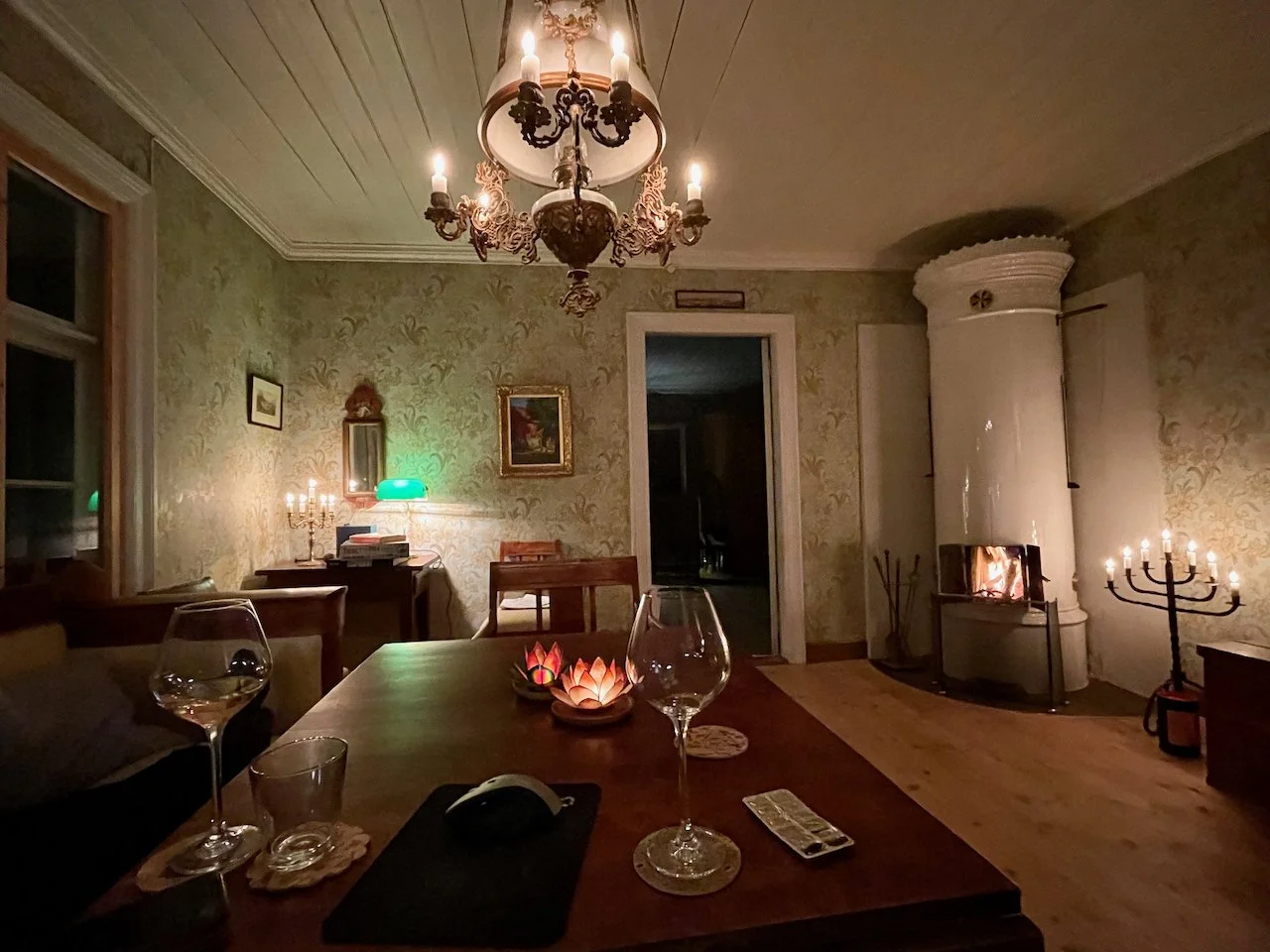 A cozy dining room with a wooden table featuring wine glasses, a censer candle, and a computer mouse. The room has vintage wallpaper, a chandelier, a fireplace, and various lamps creating a warm ambiance.
