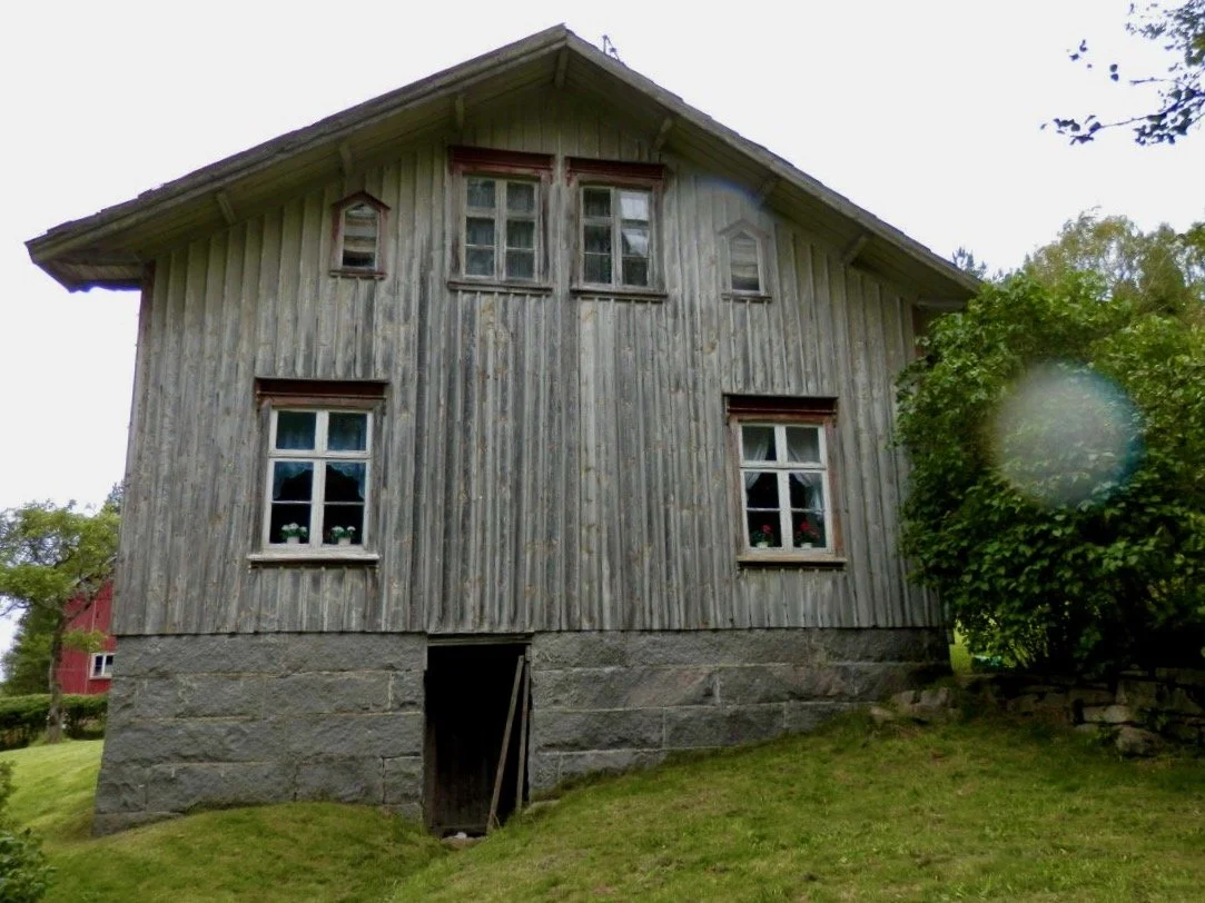 An old wooden house with a stone foundation, four windows on the front, and a small door at the bottom. The house has a slanted roof and is surrounded by green grass and trees.