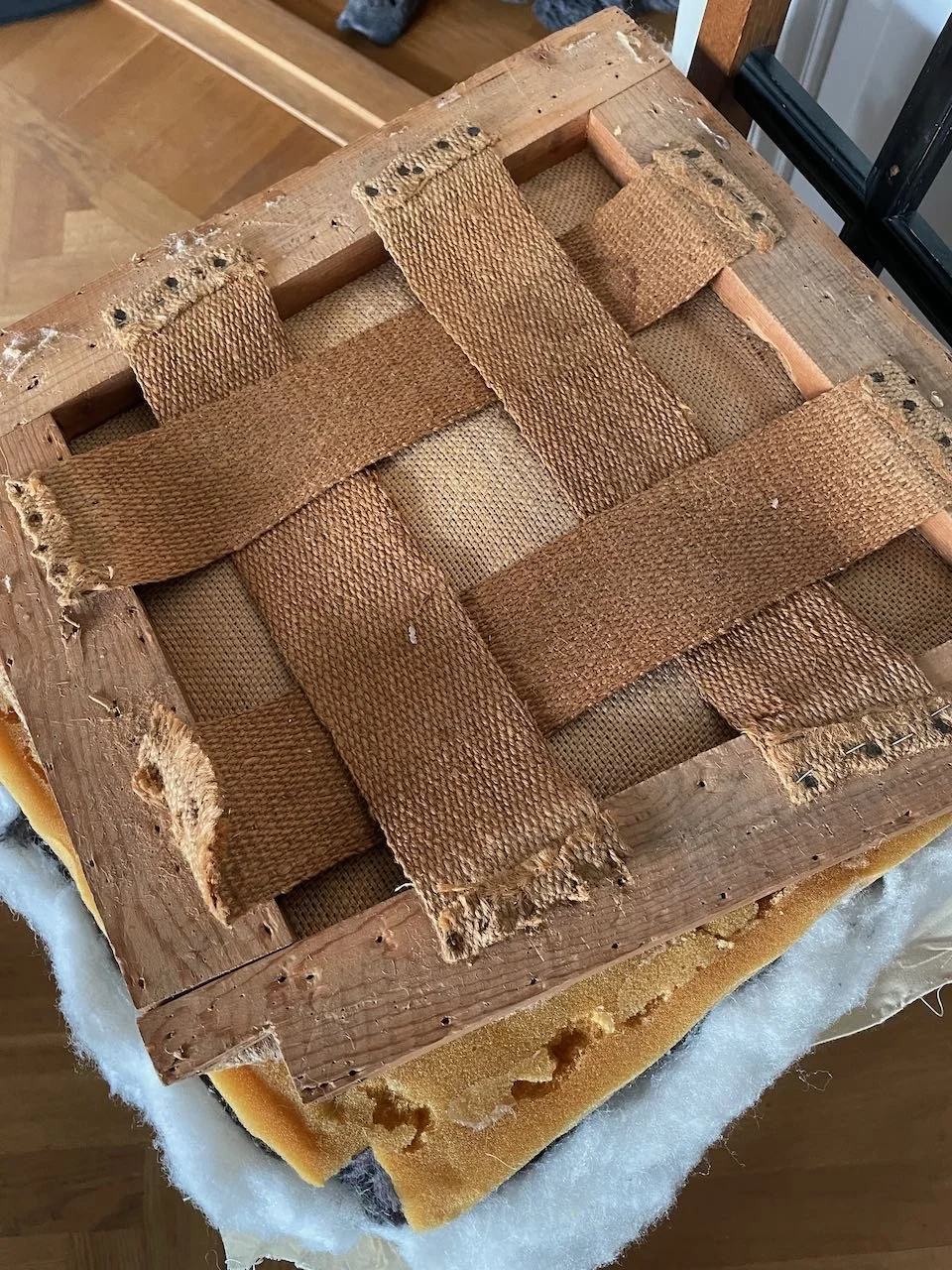 Close-up of a wooden chair with damaged woven seat, showing frayed and torn strips of woven material, likely burlap or similar fabric, with upholstery foam visible underneath.