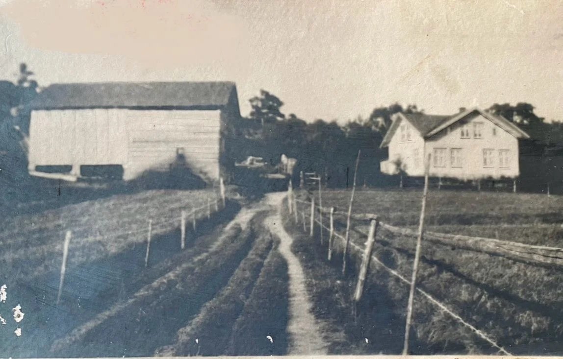 A black-and-white photograph of a rural scene with a dirt road leading to a house and a barn in the background. The road is flanked by a simple wire fence on both sides.
