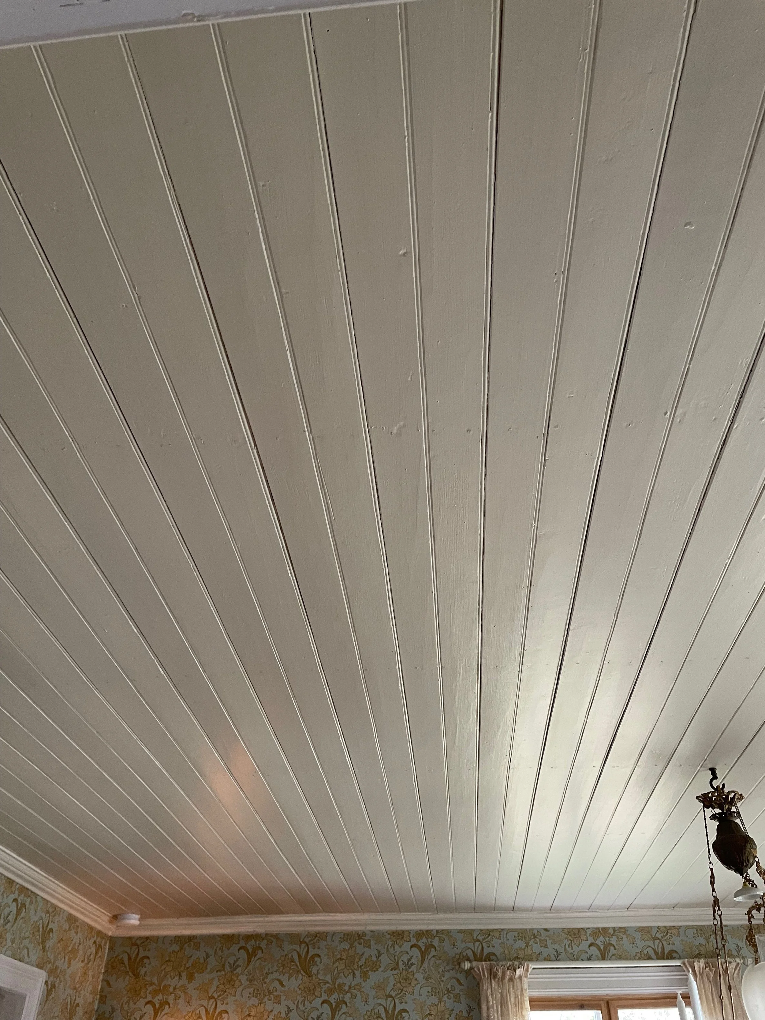 Interior view of a room with a wood-paneled ceiling painted beige, floral wallpaper on the walls, and a window with curtains. An ornate ceiling lamp with a chain is also visible.