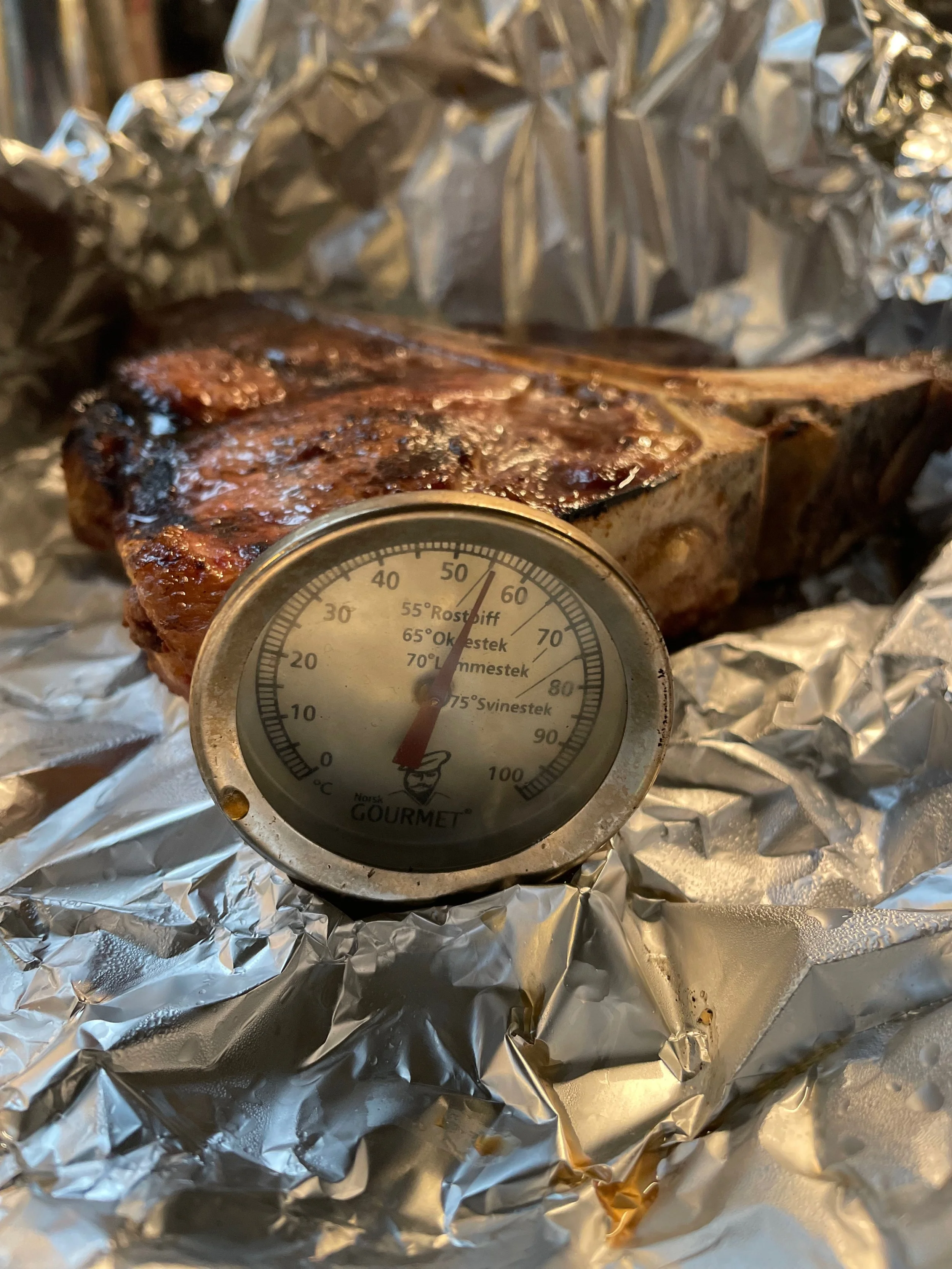 T-bone steak resting in aluminum foil with a meat thermometer showing 61°C.