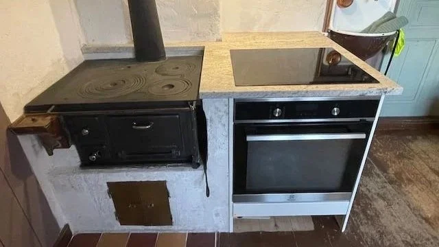 Cooking area with a black wood stove and a modern black and silver oven, separated by a granite countertop, in a kitchen with wood floor and tiles infront of wood stove.