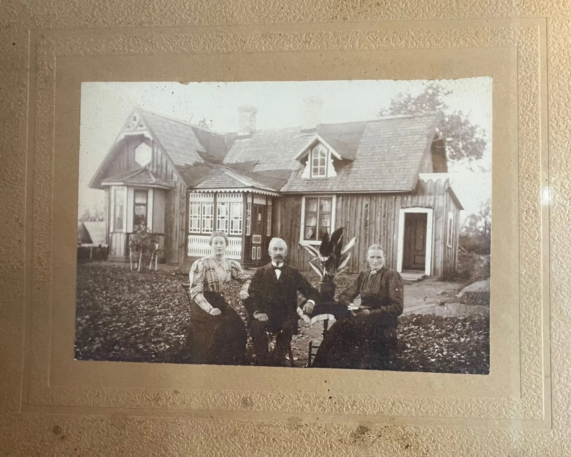 Black and white photograph of a house with several gabled roofs and a porch. Three people, a woman and two men, are sitting in front of the house.