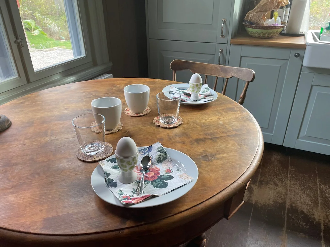 A wooden dining table set for breakfast with two plates, each holding a soft-boiled egg in an egg cup, a spoon, a napkin, and a glass of water. There are also two mugs, two glasses, and coasters on the table. The background shows a window with a view