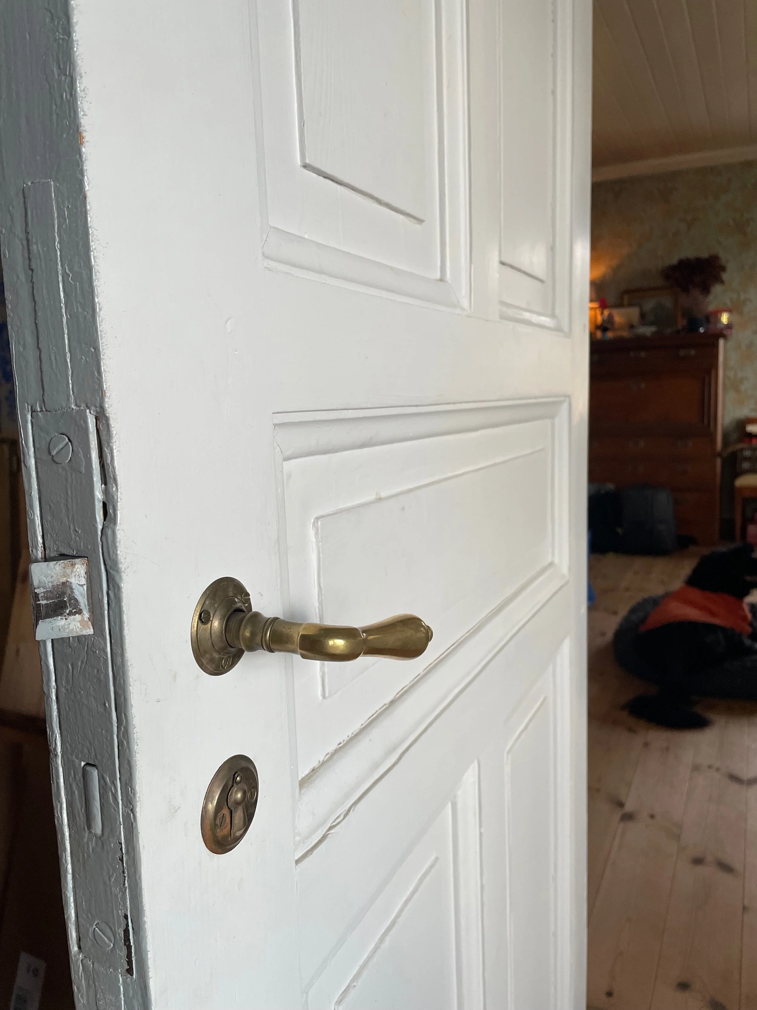 Close-up of a white door with brass door handle and lock, slightly open, showing a room inside with a wooden dresser and various items on top, and a brick floor.