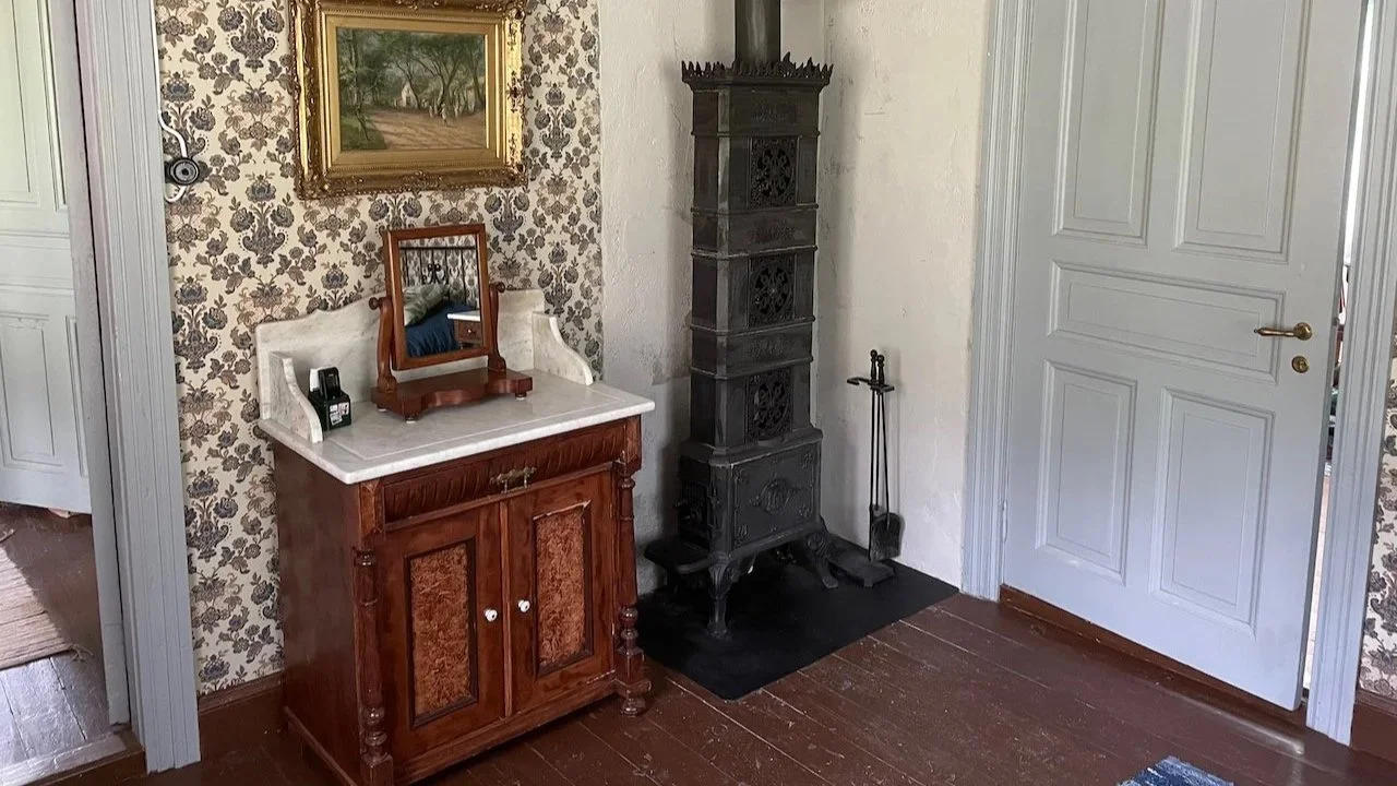A vintage interior corner with a wooden cabinet topped with a small mirror and a landline telephone, an ornate black cast iron stove, a pair of fireplace tools, and a framed landscape painting on wallpapered wall next to a white door.
