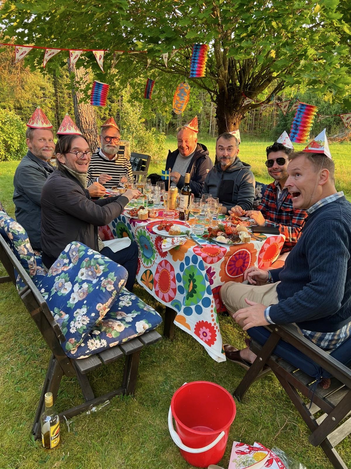 Group of seven friends having the yearly cray-fish party, wearing colorful party hats, seated at a decorated table with food and drinks, under a tree with festive decorations, in a backyard with green grass and trees.