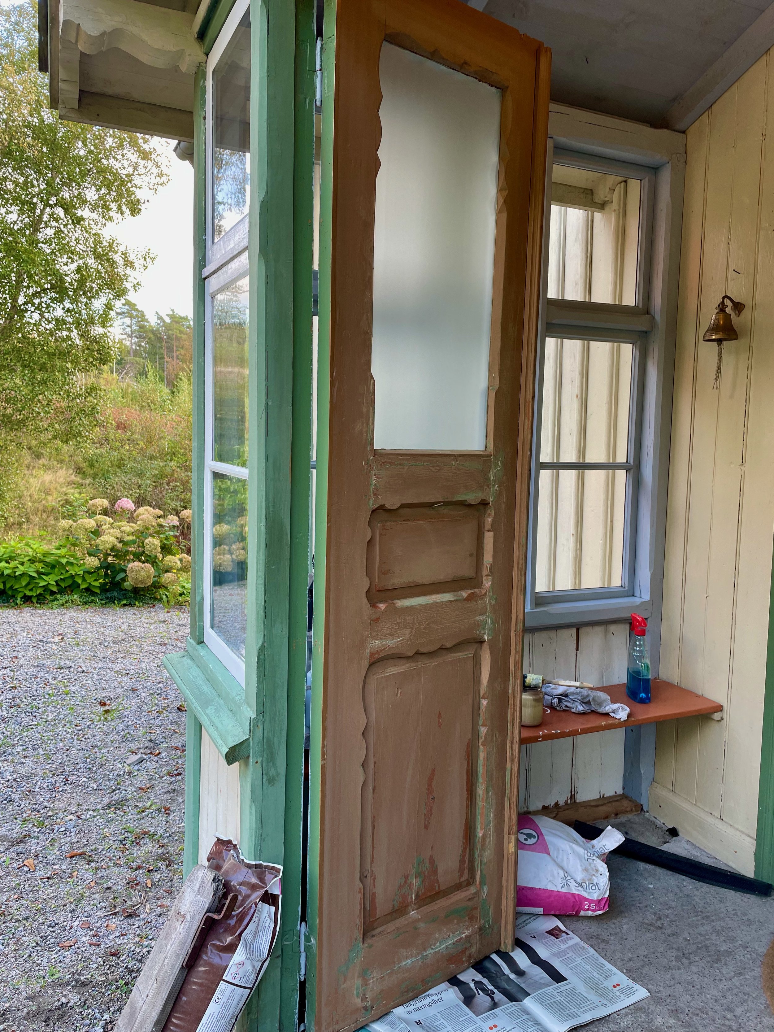 A partially painted wooden open door of a small porch with a garden outside, and some cleaning supplies on a small ledge inside.