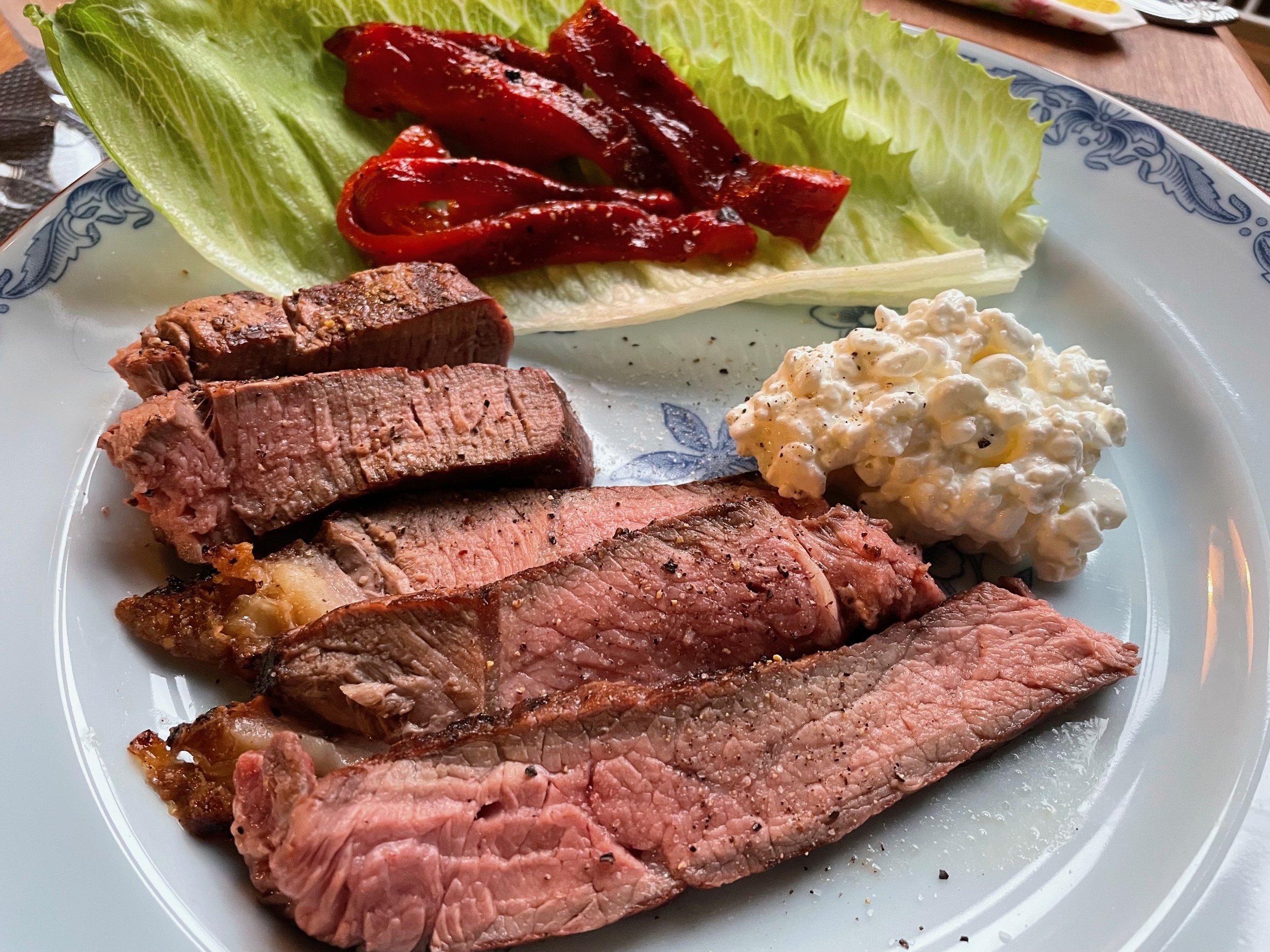 A plate of cooked steak with slices arranged on a white and blue decorative plate. Accompanying the steak are leafy Romaine lettuce and red bell pepper strips, and a dollop of creamy cottage cheese or potato salad. The background shows a wooden table and some utensils.