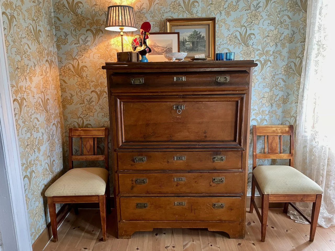 A vintage wooden dresser with five drawers and a cabinet, flanked by two wooden chairs with upholstered seats, inside a room with floral wallpaper and lace curtains.