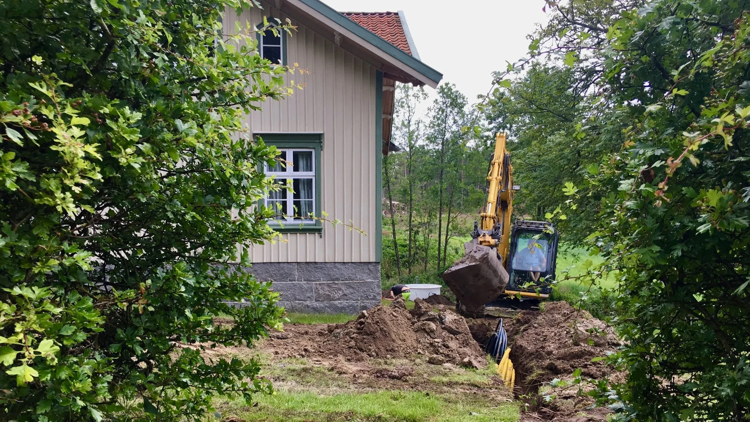 A construction site next to a beige house with green trim, showing a yellow excavator digging a trench in the ground surrounded by green trees and bushes.