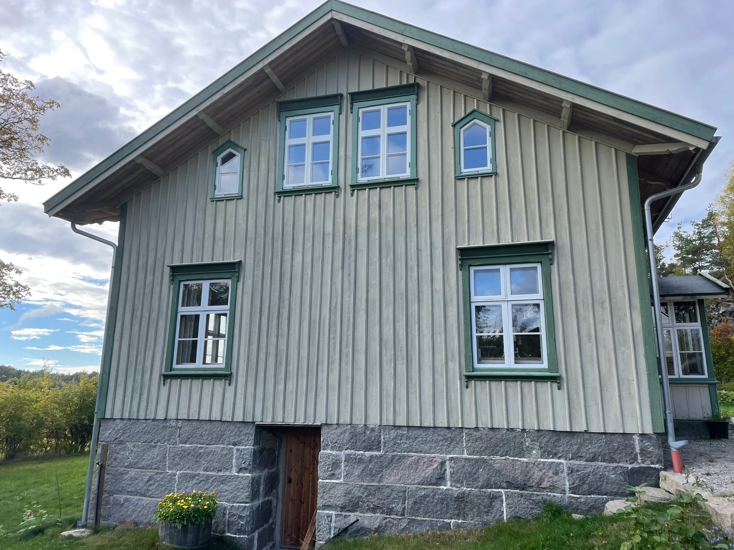 A two-story traditional house with wooden siding, multiple windows with green frames, and a stone foundation, set against a partly cloudy sky with greenery around.