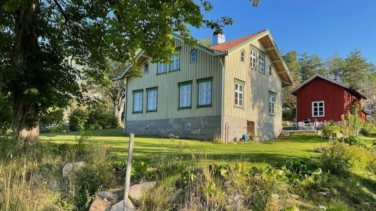 A sand colored house with a stone foundation and multiple windows, surrounded by trees and a well-maintained lawn, with a smaller red building nearby and outdoor furniture under a clear blue sky.