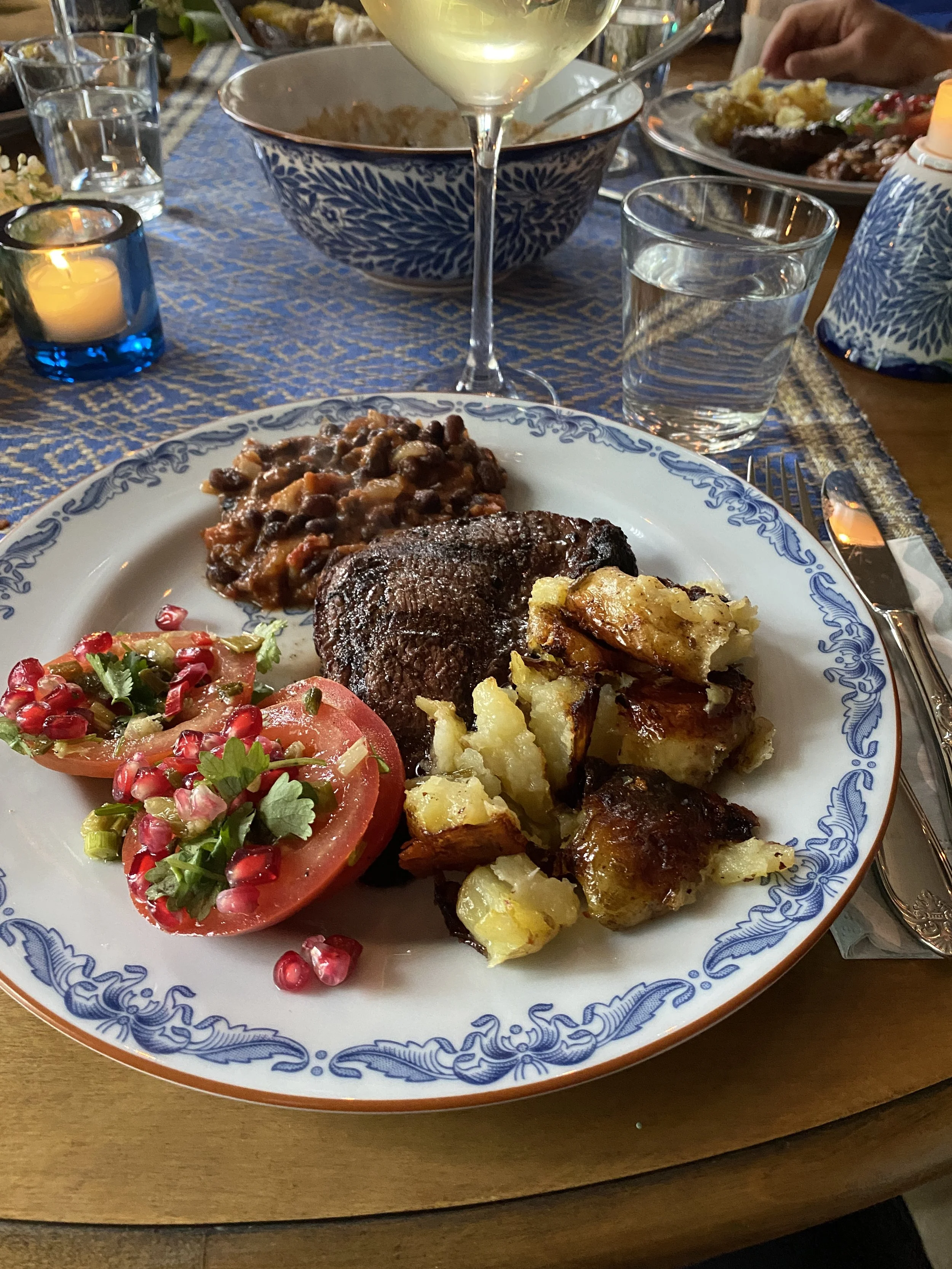 A plate of food with baked potato, grilled beef steak, tomato and pomegranate salad, and a serving of beans or lentils. In the background, a glass of white wine, a glass of water, and other dishes and utensils on a dining table.