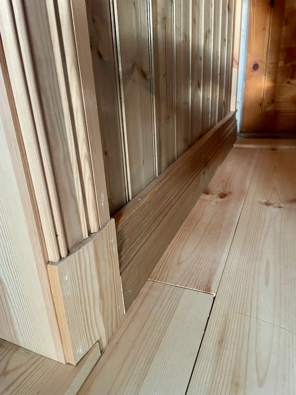 Close-up photo of a wooden baseboard along a wall with vertical wooden paneling and horizontal wooden flooring.
