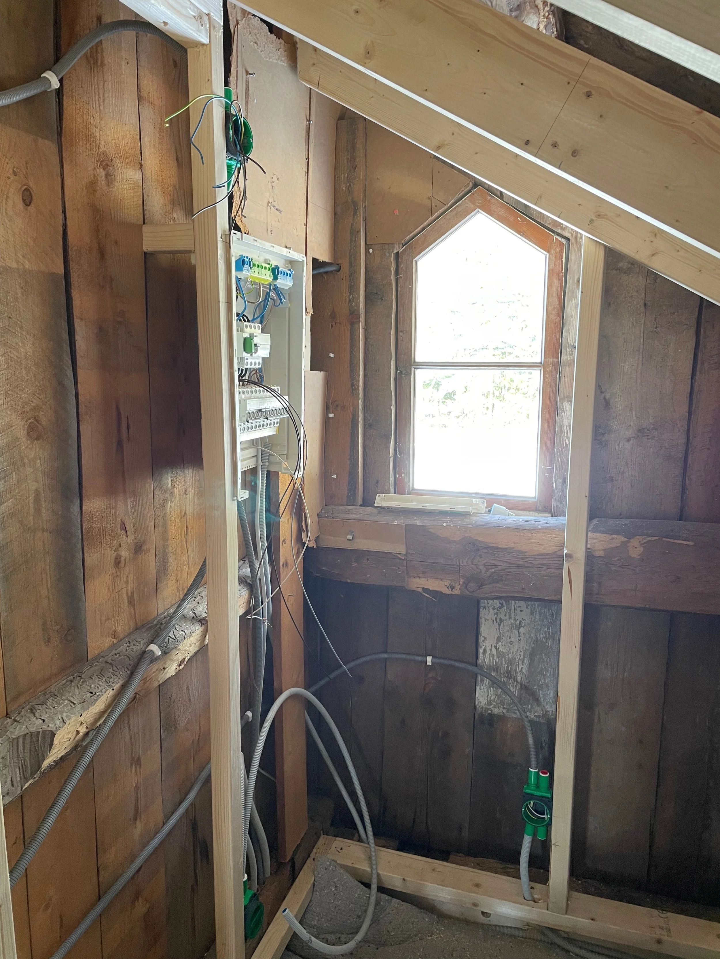 Interior of a building under construction or renovation, with exposed wooden framing, electrical wiring, a control box, and a window.