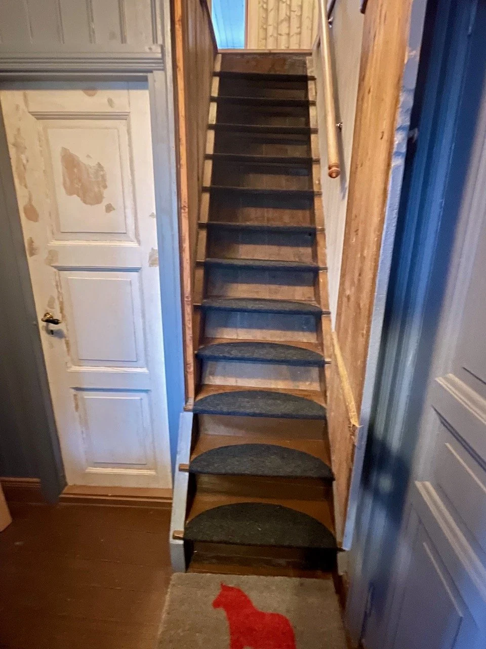 A narrow wooden staircase with black carpeted steps leading upwards, with a wooden handrail on the right side. To the left, there is a white door with peeling paint and a brass handle. A small rug with a red silhouette of a cat is on the floor at the bottom of the stairs.