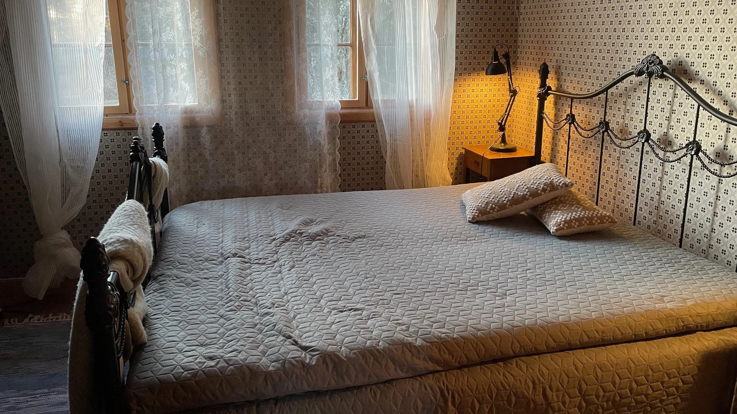 A bedroom with a metal-framed bed, quilted mattress, and two pillows, with light coming through lace curtains on the windows, and a yellow wall with patterned wallpaper, a nightstand, and a lamp.