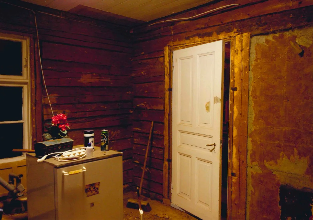 Interior of a room under renovation with exposed wooden walls, a white door, a window, and a small cabinet with household items on top.