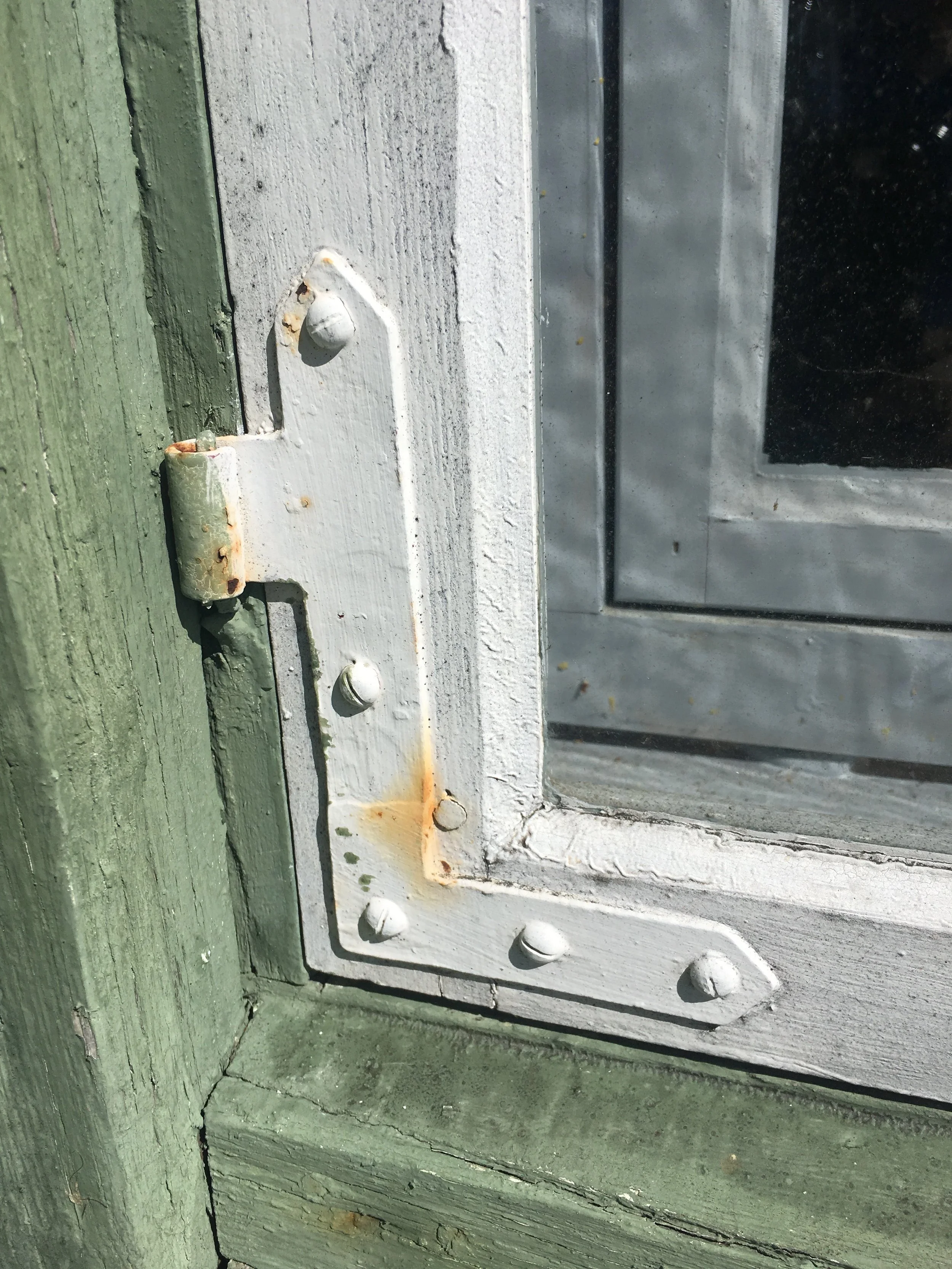 Close-up of a white-painted wooden window frame with visible rust, chipping paint, and rusted screws, set in a green wooden window trim frame.