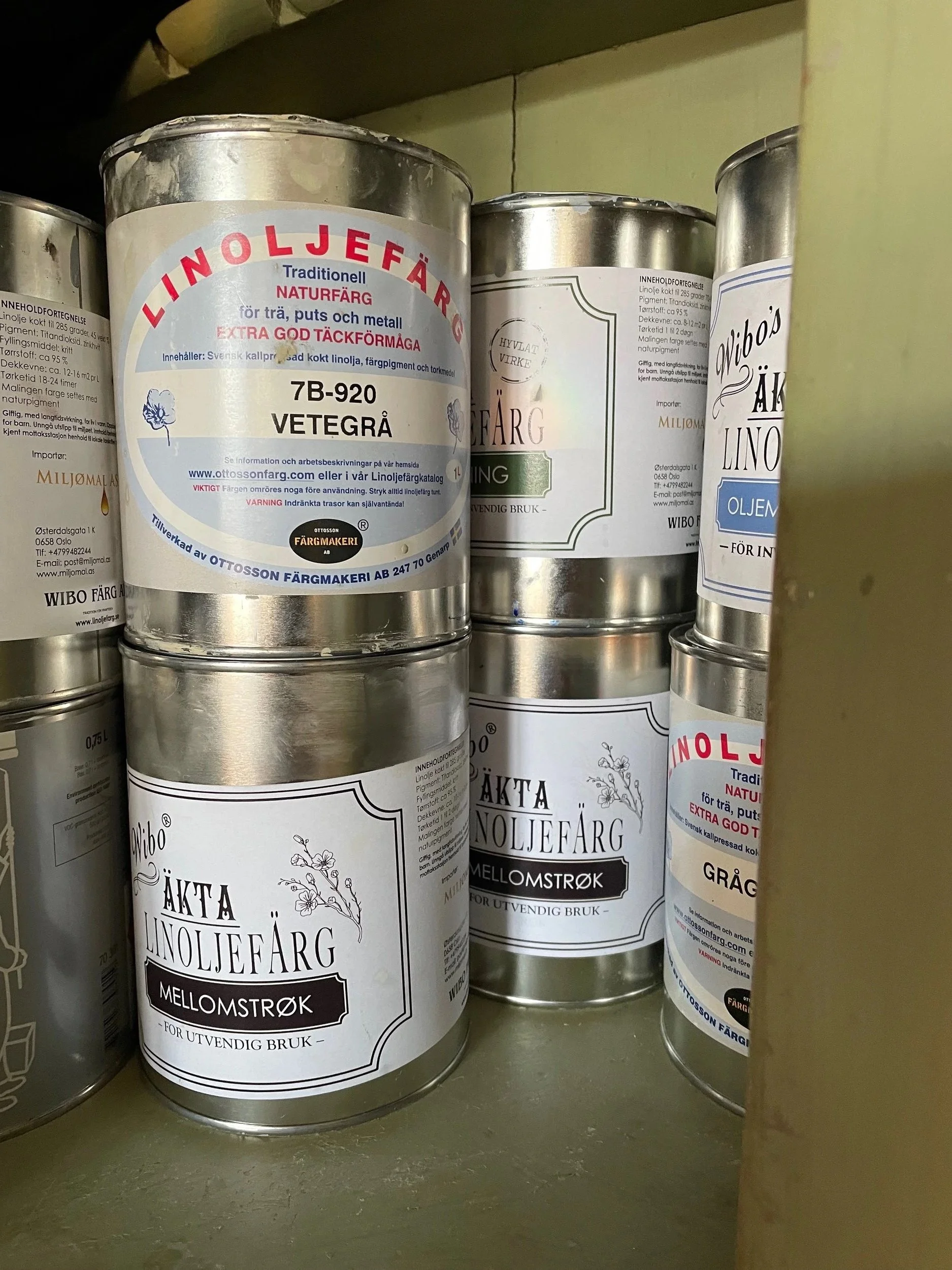 Paint cans on a shelf, labeled with Swedish text indicating different linseed oil paint colors and types, including linoleum paint in medium shade.