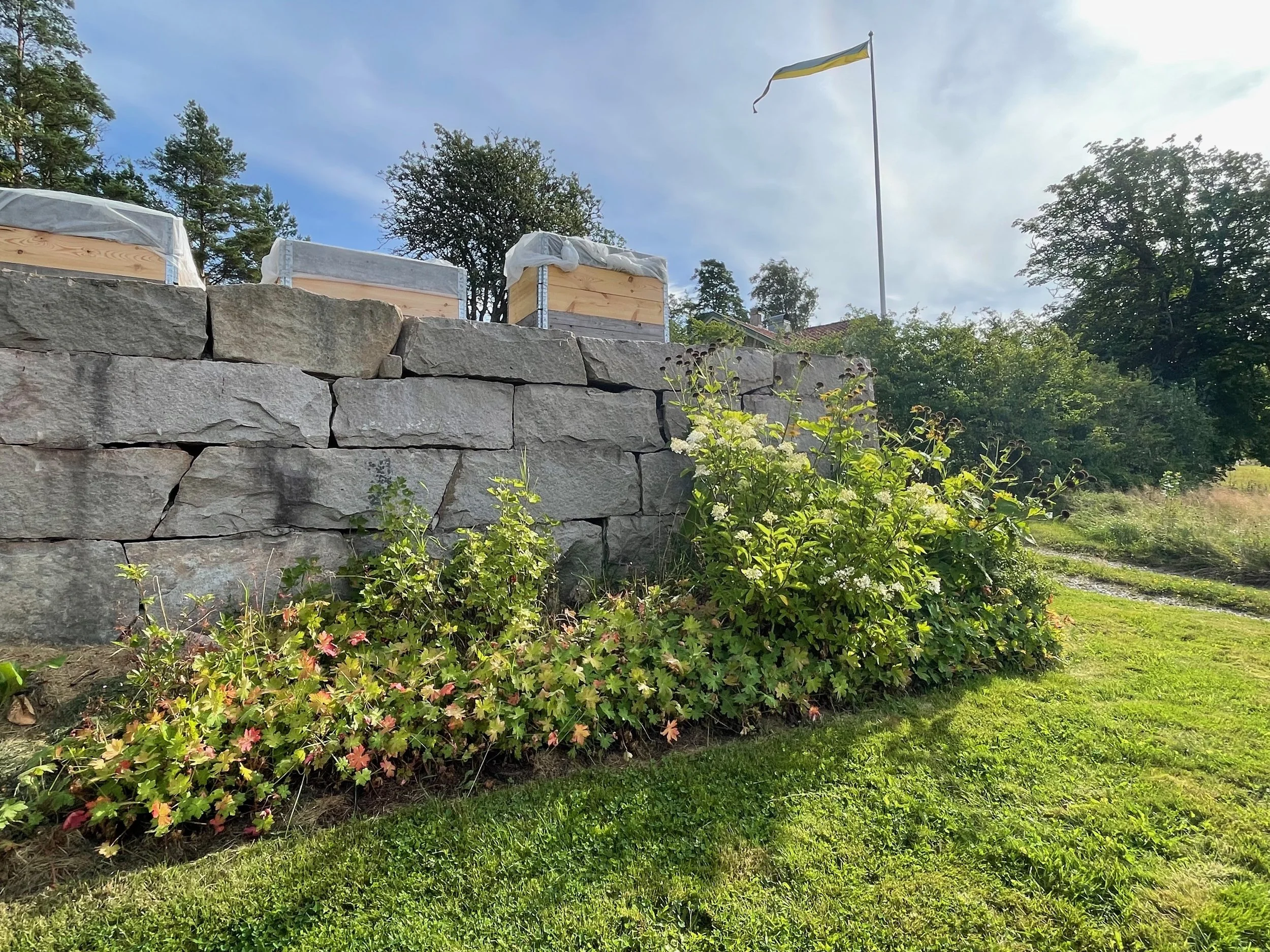 A stone garden wall with wooden boxes on top, a yellow and grey flag flying in the background, trees, and flowering bushes in the foreground, with a partly cloudy sky.