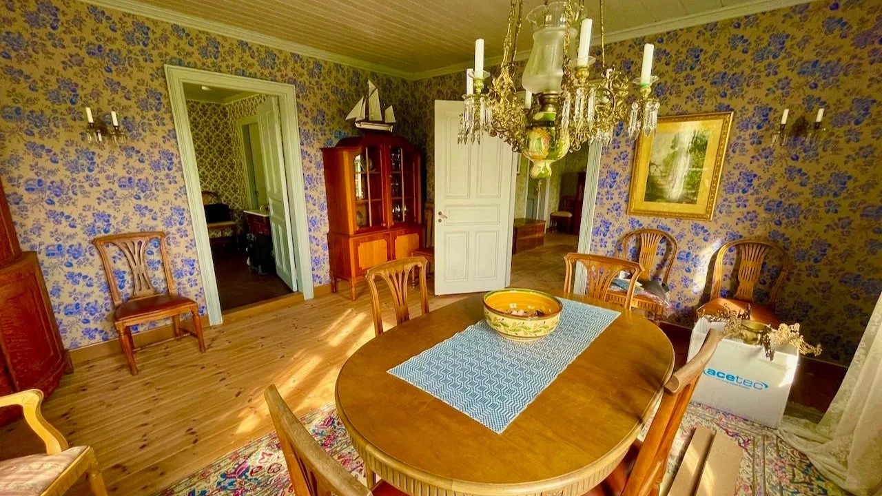 A dining room with floral wallpaper, a wooden dining table with six chairs, a blue table runner, a ceramic bowl, chandelier, wall-mounted candle sconces, a painting, wooden cabinets, an open door leading to another room, and a white box on the floor.