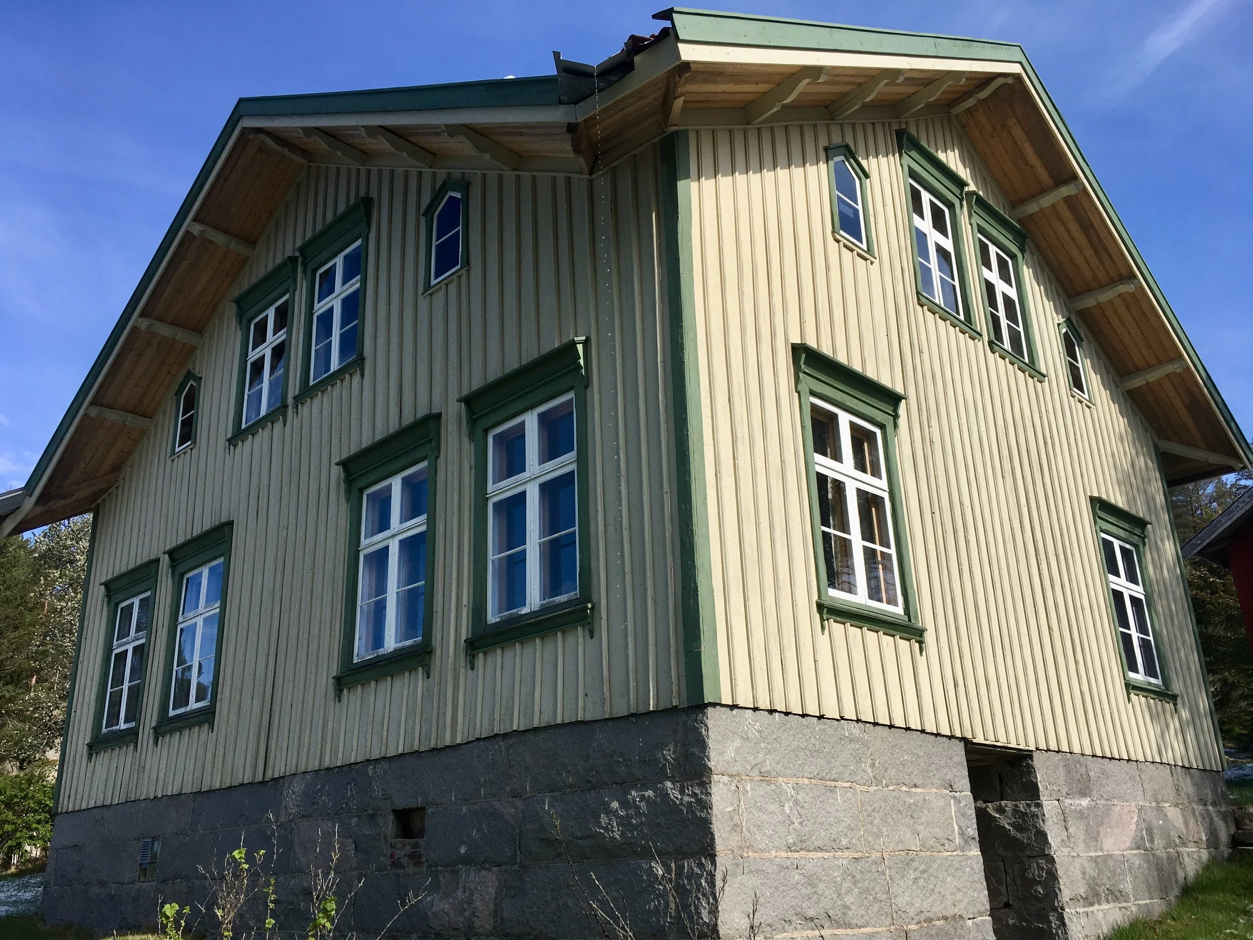 A two-story house with a green exterior and white window frames, built on a stone foundation, under a blue sky.