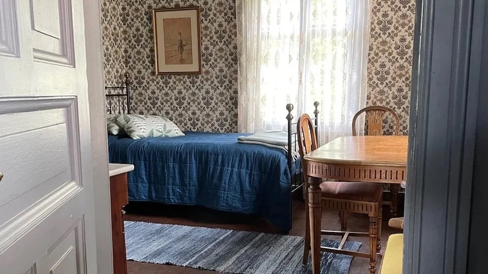 A cozy bedroom with floral wallpaper, a bed with a blue quilt, pillows, and a framed art piece on the wall. There is a wooden dining table with chairs near a window with sheer curtains.