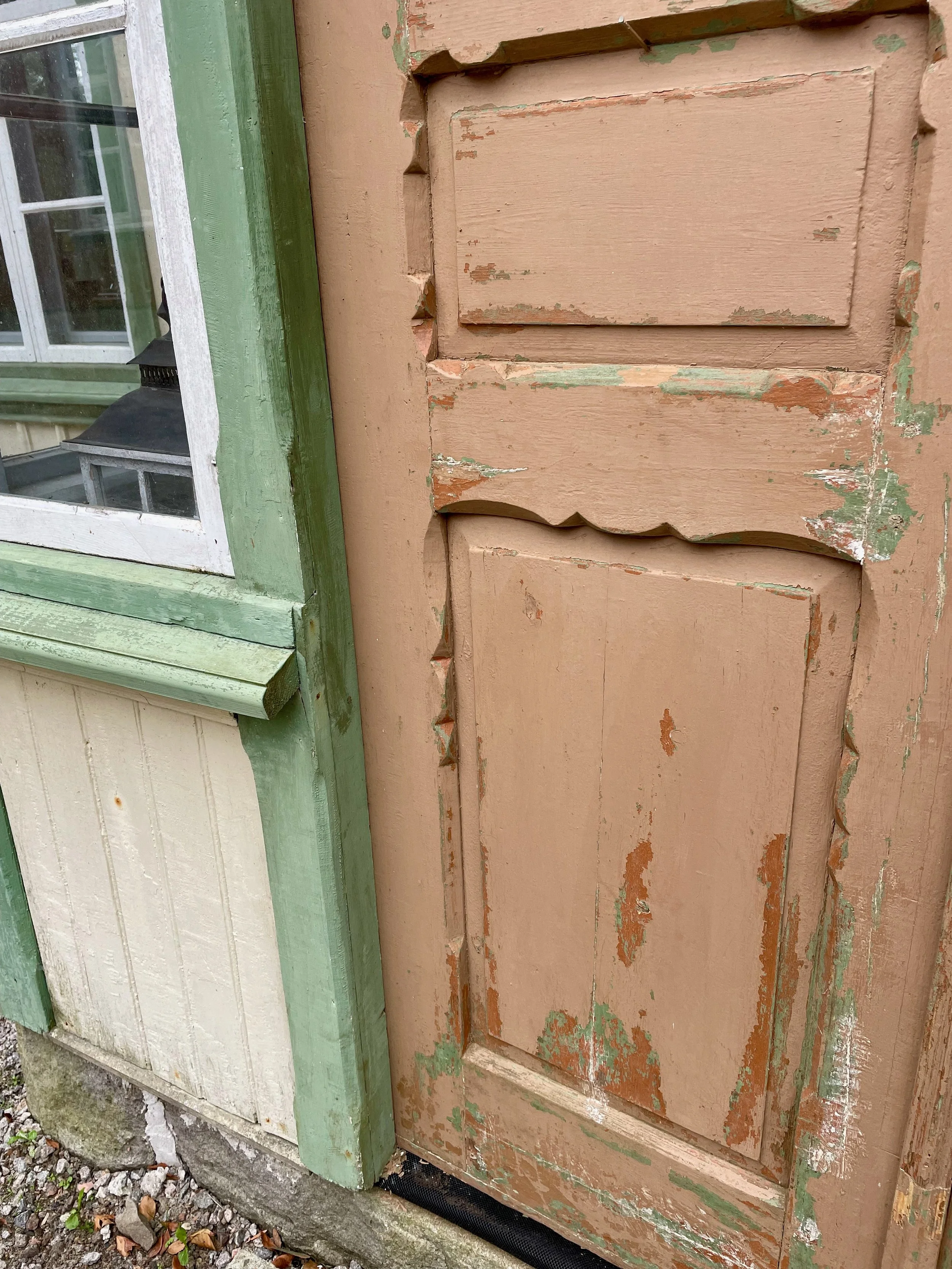 Close-up of a weathered, peeling wooden door with painted decorative panels, next to a window with green trim on a weathered house.