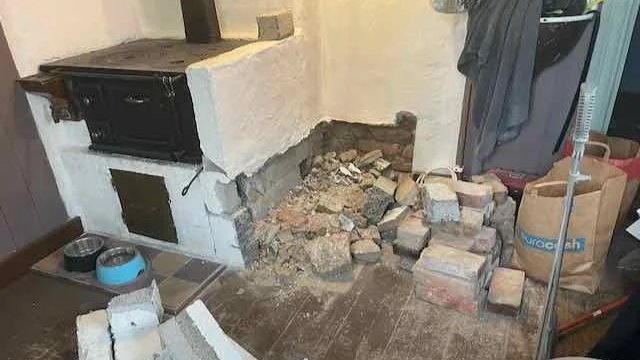 Disorganized pile of bricks and rubble next to a white wall with an old black stove, on a wooden floor.